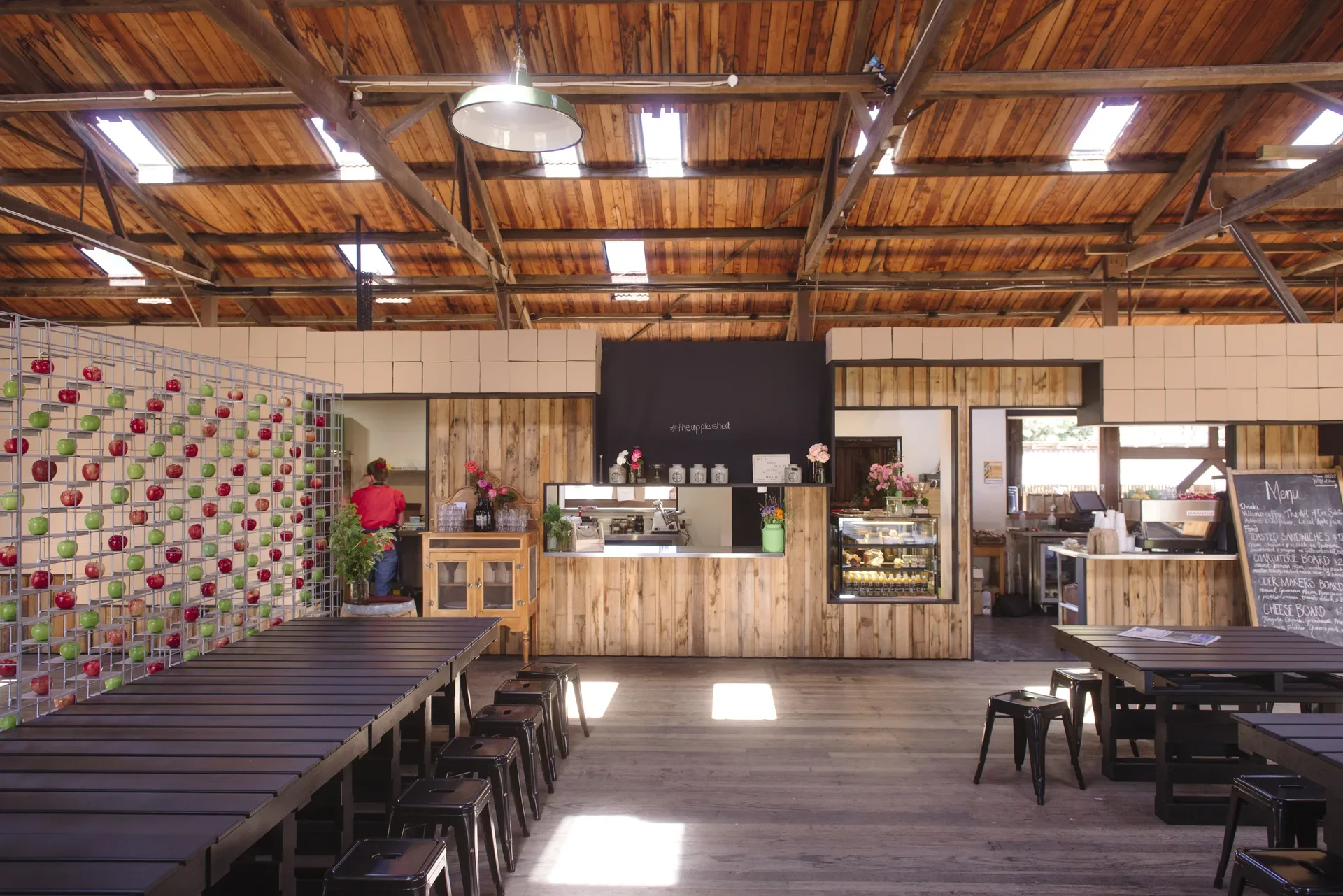 This rustic interior features a vaulted timber ceiling with exposed beams and skylights above long black communal tables. In the background, a service counter clad in vertical wood planks and topped with cardboard boxes sits next to a large wire grid wall displaying dozens of red and green apples.