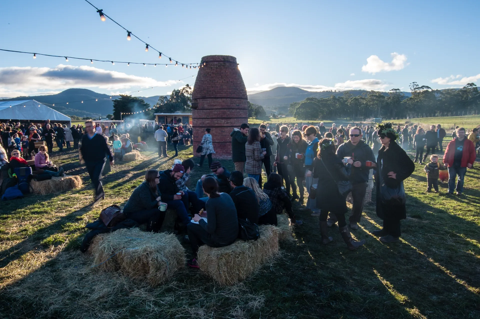 This outdoor event features a large crowd gathered on a grassy field under a clear blue sky. People are socializing near a tall brick kiln-like structure and sitting on hay bales, while strings of outdoor lights hang overhead.