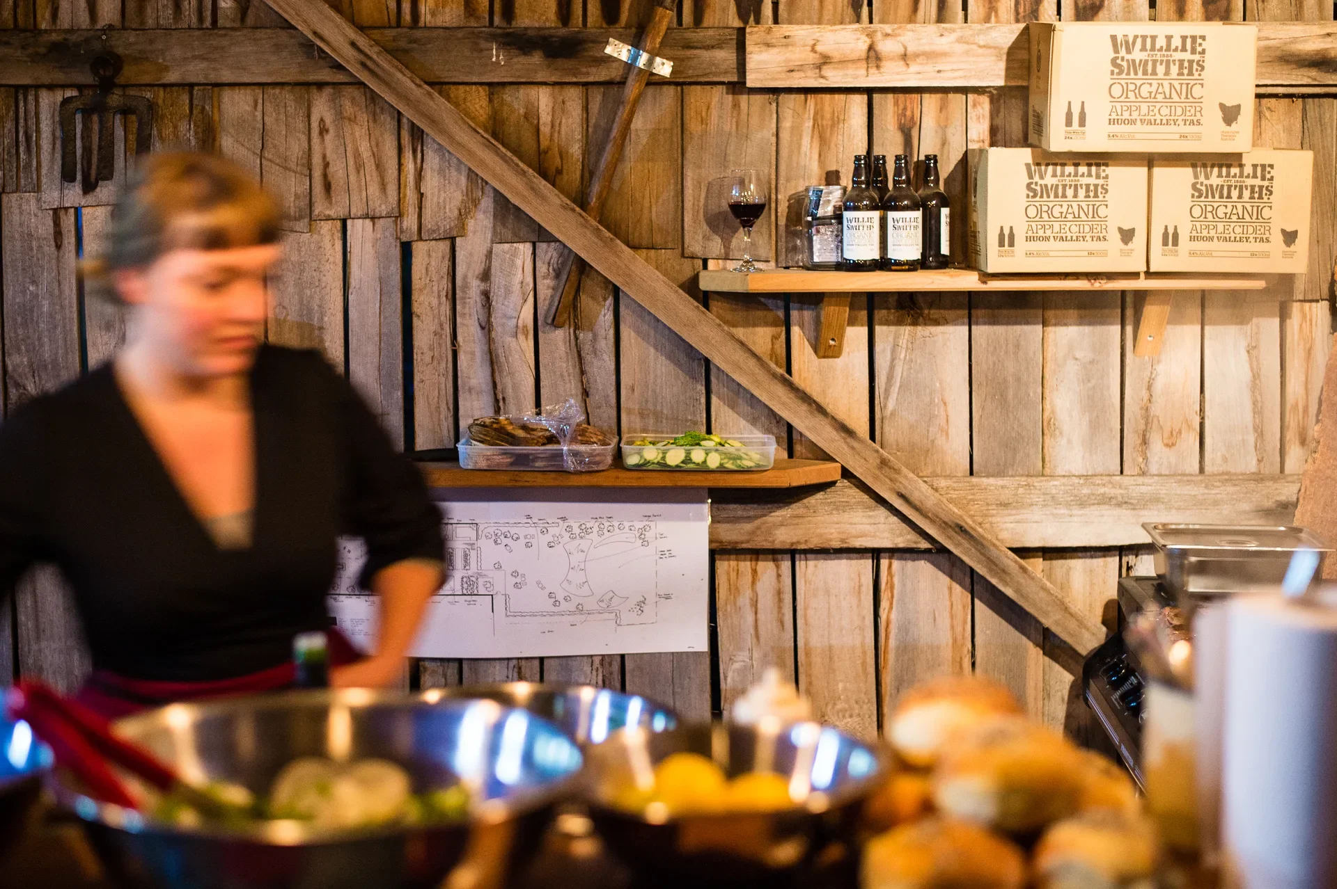 This busy kitchen features vertical timber walls. A wooden shelf holds Willie Smith’s Organic Apple Cider bottles and branded shipping boxes, while fresh ingredients and metal mixing bowls fill the foreground.