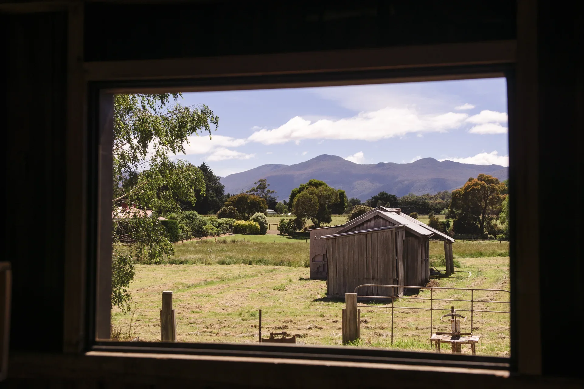 This framed view through a window reveals a rustic landscape of green fields and a weathered wooden shed. In the distance, blue-tinted mountains sit under a bright sky with scattered white clouds.