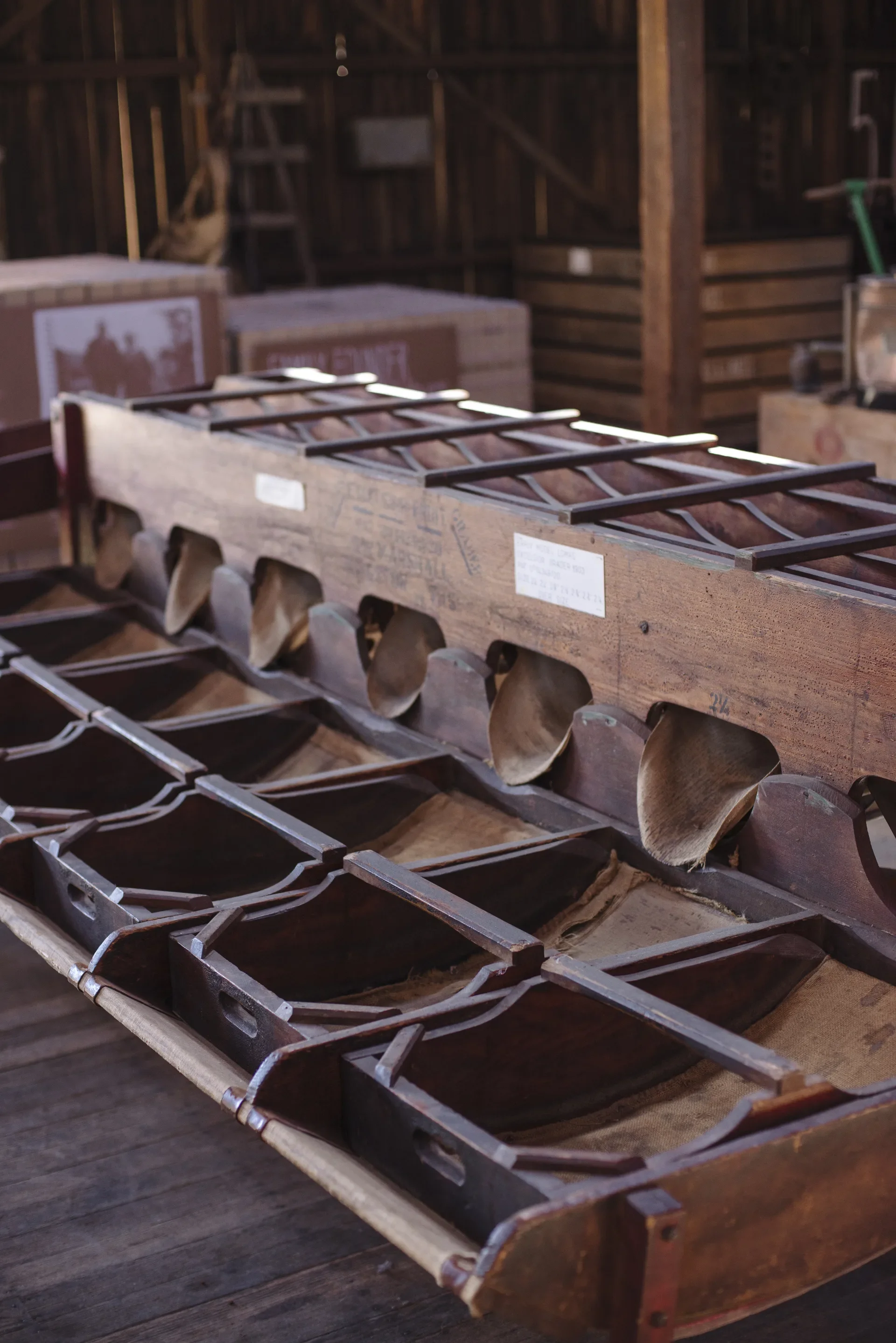 This museum display features a large vintage wooden apple grader with several collection bins lined with burlap. The hand-crafted machine is set within a rustic timber shed, with stacked wooden crates visible in the background.