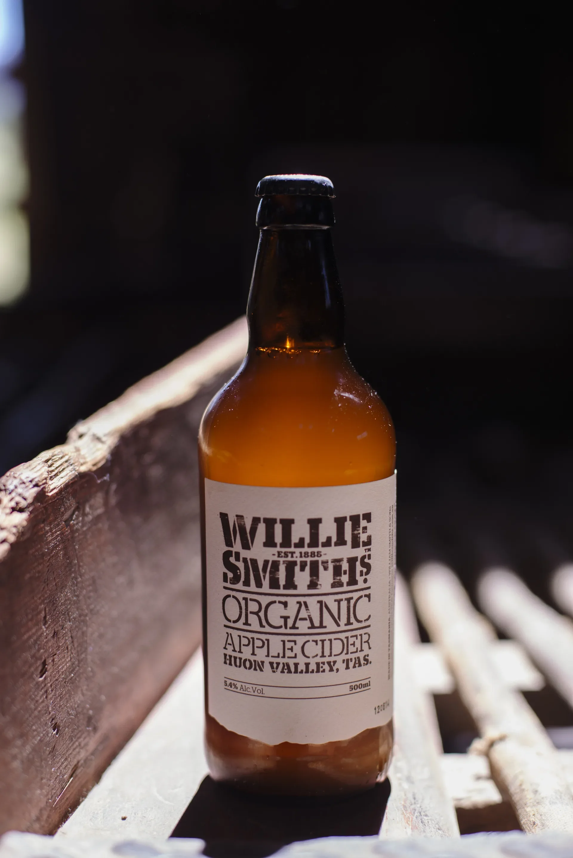 A bottle of Willie Smith’s Organic Apple Cider sits on a weathered wooden surface under bright day light. The rustic label clearly displays the Huon Valley, Tasmania origin against a dark, shadowed background.