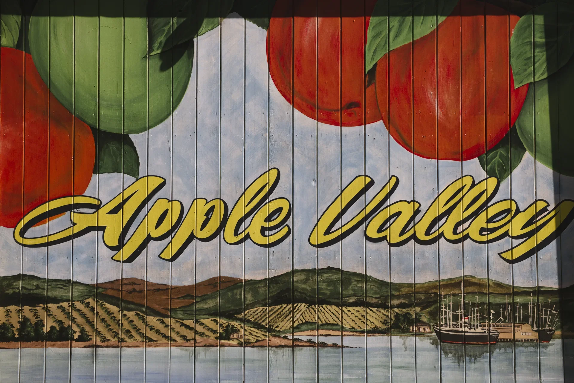 This vibrant "Apple Valley" mural is painted on a wall of vertical timber slats. It features large red and green apples at the top, with a scenic landscape of rolling hills, orchards, and a sailing ship in a harbour below.