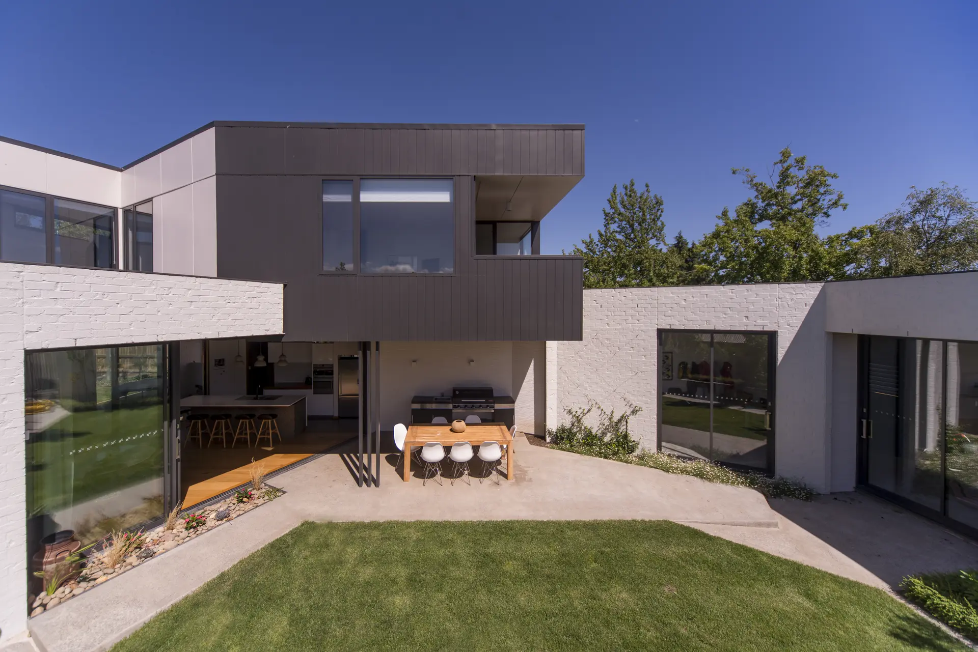 A photograph of the rear elevation showing the house opening to a central courtyard, with white-painted brick volumes at ground level and a dark, vertically clad upper form hovering above. Full-height sliding glazing connects living spaces to a shaded outdoor dining area and lawn.