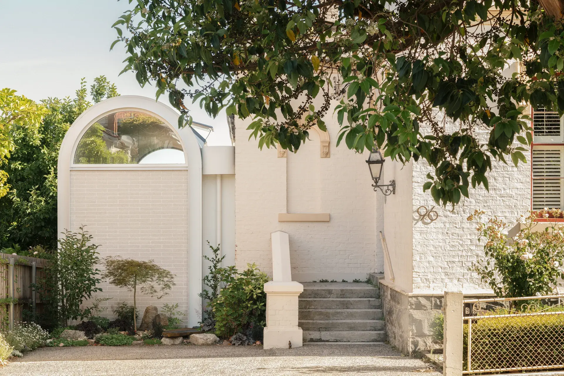 This light-coloured masonry building features a prominent arched window and a short flight of concrete stairs leading to an entrance. The exterior is decorated with a vintage-style black lantern, while lush greenery and the branches of a leafy tree frame the scene.