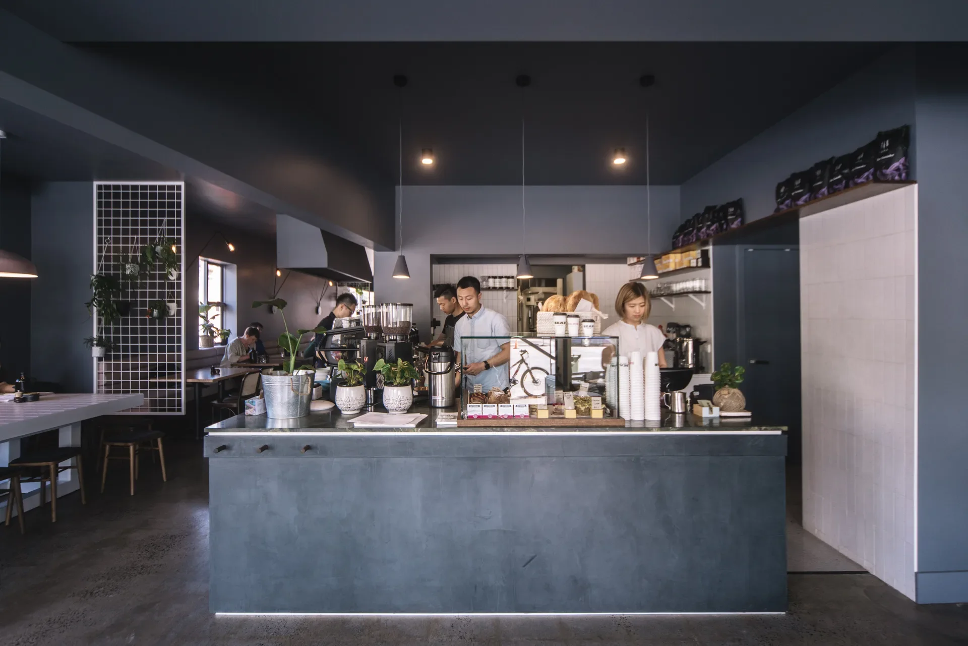 A monochrome space uses dark grey, white accents, and strategic lighting to create a high-contrast backdrop for a front counter occupied by staff and filled with plants and bakery goods.