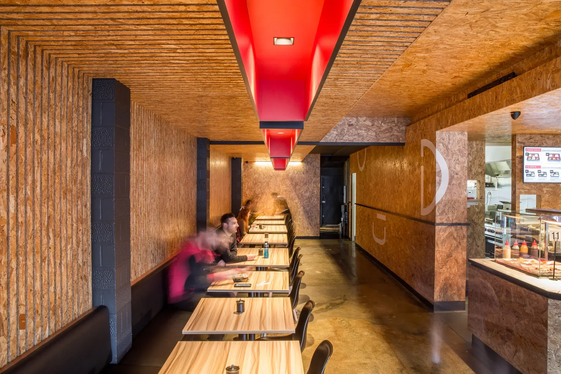 A narrow dining space at Bento Moonah is lined entirely with textured OSB wall panels and vertical timber battens, with a continuous red-lit ceiling element drawing the eye through the length of the room. Simple timber tables and bench seating sit beneath warm, ambient lighting, creating an intimate, linear dining experience.