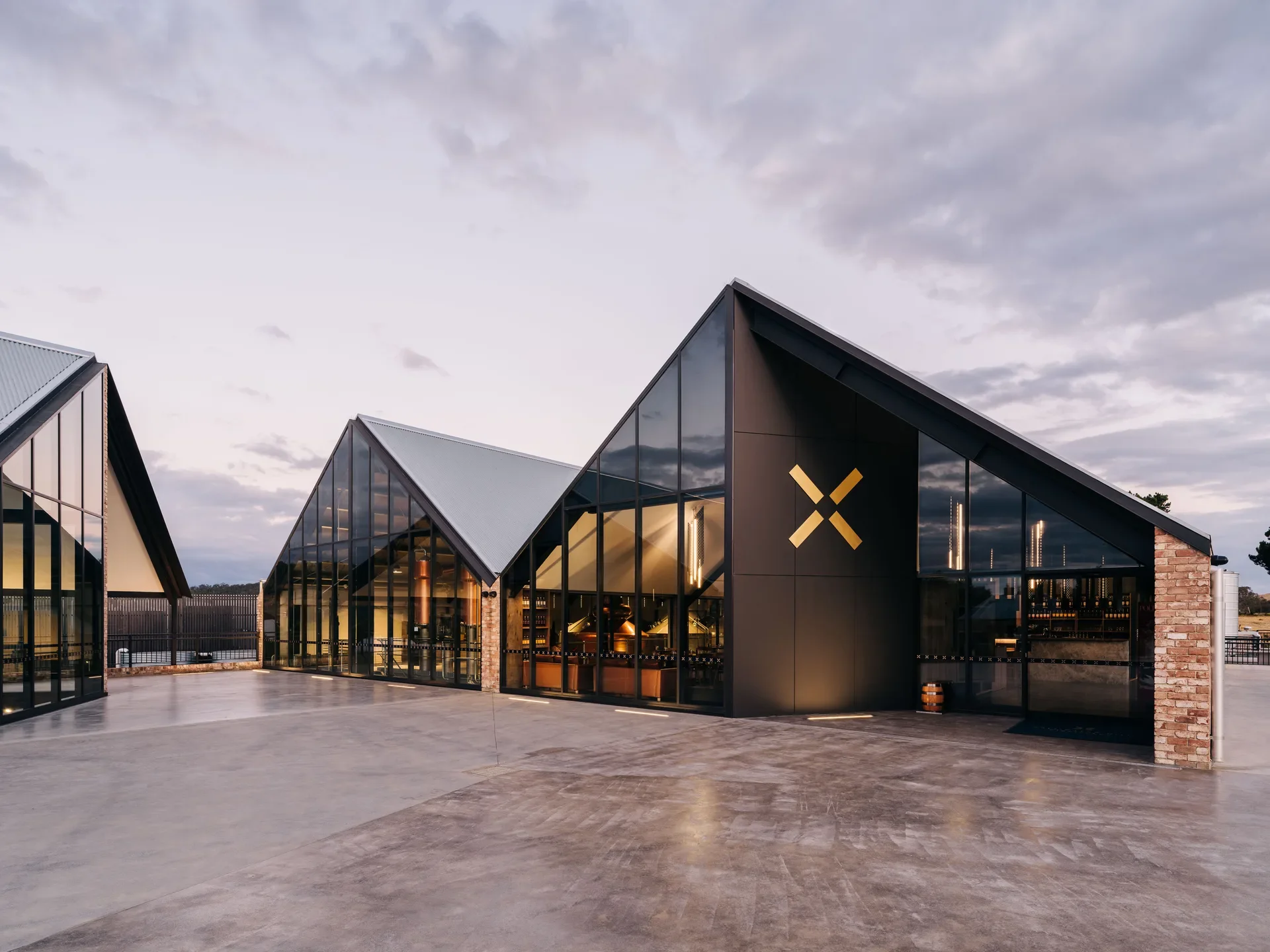 A series of black, gable-roofed buildings with expansive glass facades and brick accents surround a polished concrete courtyard, featuring a prominent yet abstract gold "X" logo representing the sails of Callington Mill.