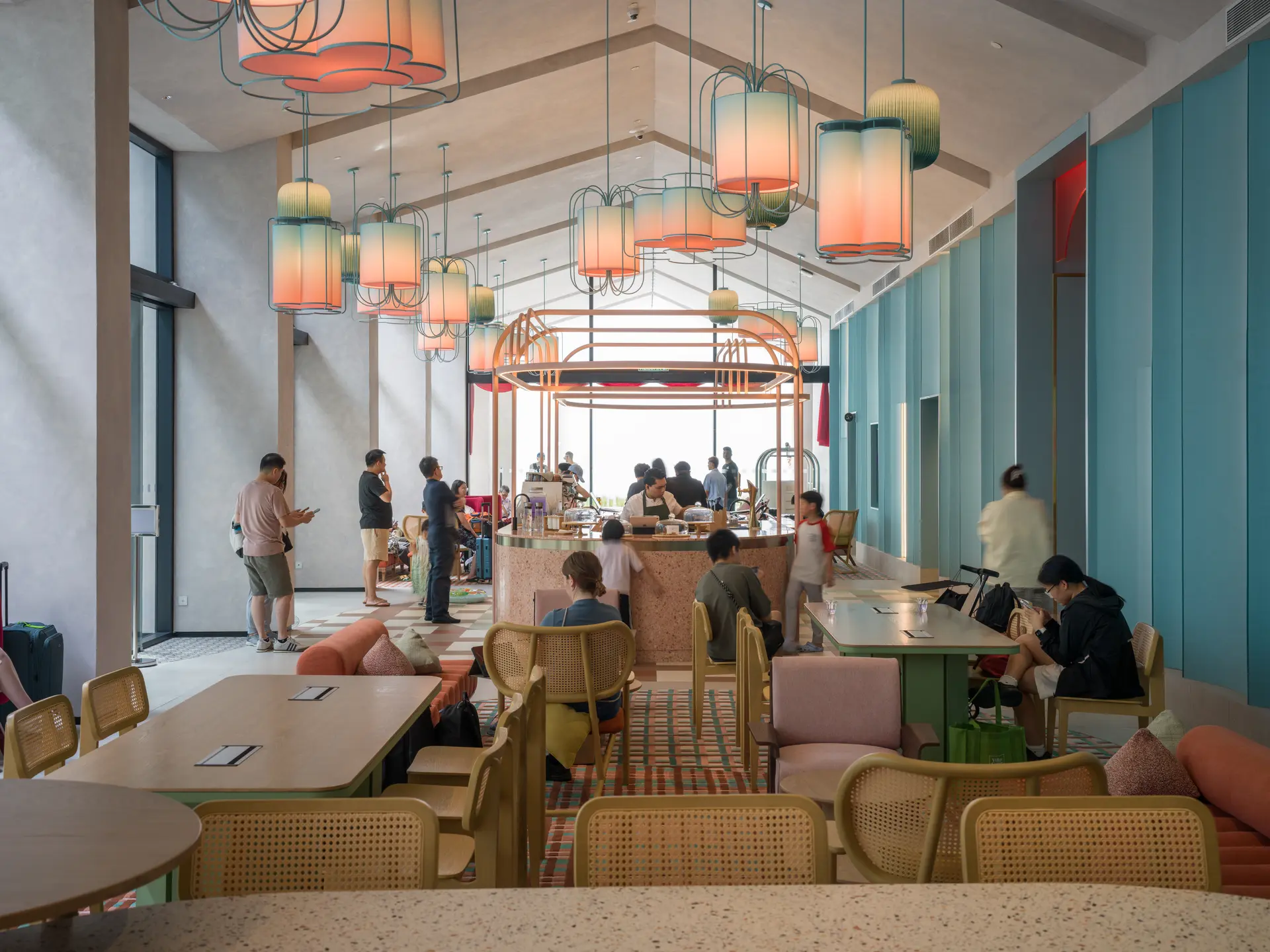 Ground-floor café with clusters of amber pendant lights over a central bar, rattan chairs and patterned rugs, and guests checking in nearby.