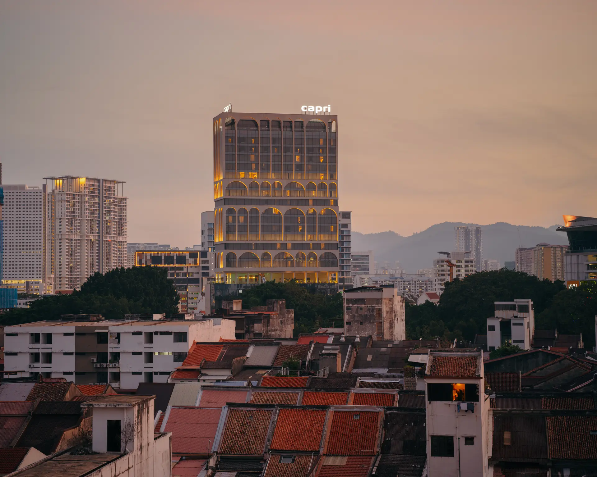 Capri Penang lit up at dusk, its arched lower floors glowing amber, standing above George Town's shophouse rooftops with hills in the distance.