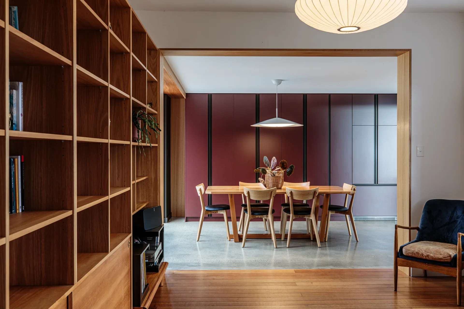 A minimalist dining room features a light timber table and chairs set against a bold, burgundy-paneled feature wall, framed by large-scale wooden shelving and warm ambient lighting.