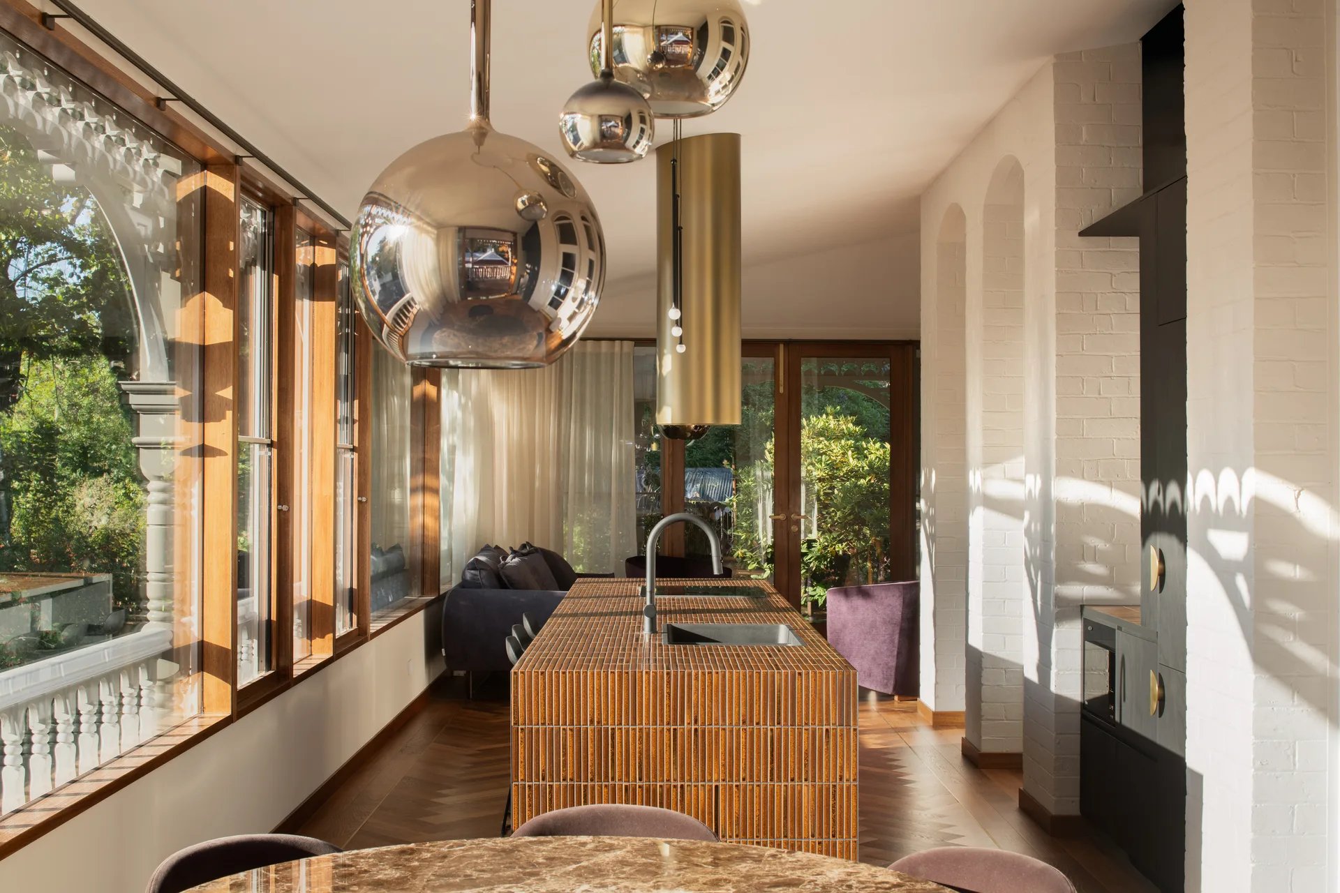 A sunlit kitchen interior with a terracotta-tiled island and integrated sink, reflective chrome globe pendants  overhead, herringbone timber flooring, and large timber-framed windows aligned with the existing verandah arches overlooking greenery.