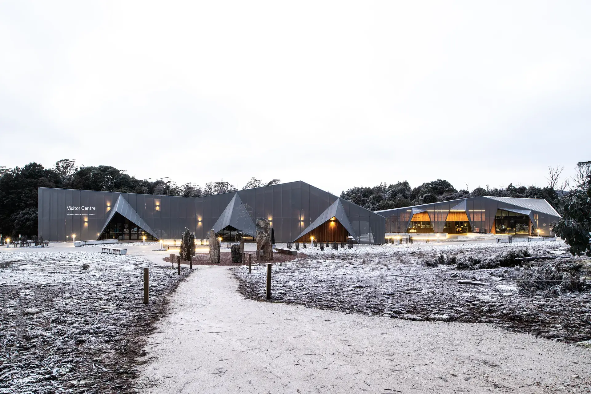 A dark, geometric visitor centre with jagged, triangular rooflines sits within a frosty landscape, its metallic facade illuminated by warm exterior lighting along a winding gravel path.