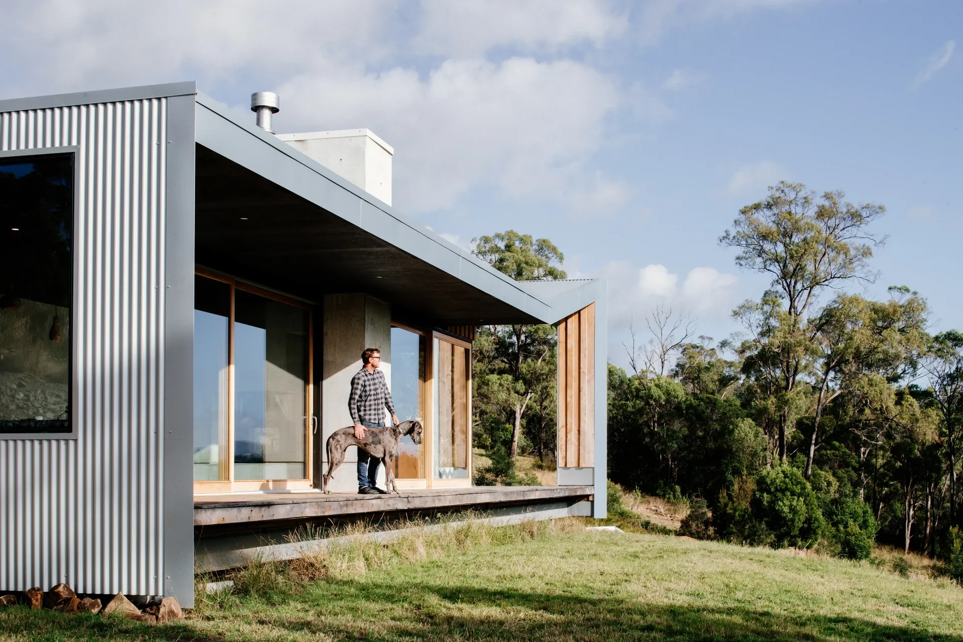 A man and his dog stand on the elevated timber deck of a modern, corrugated metal-clad home, which features a deep overhanging roof and large glass doors that open toward a grassy hillside.