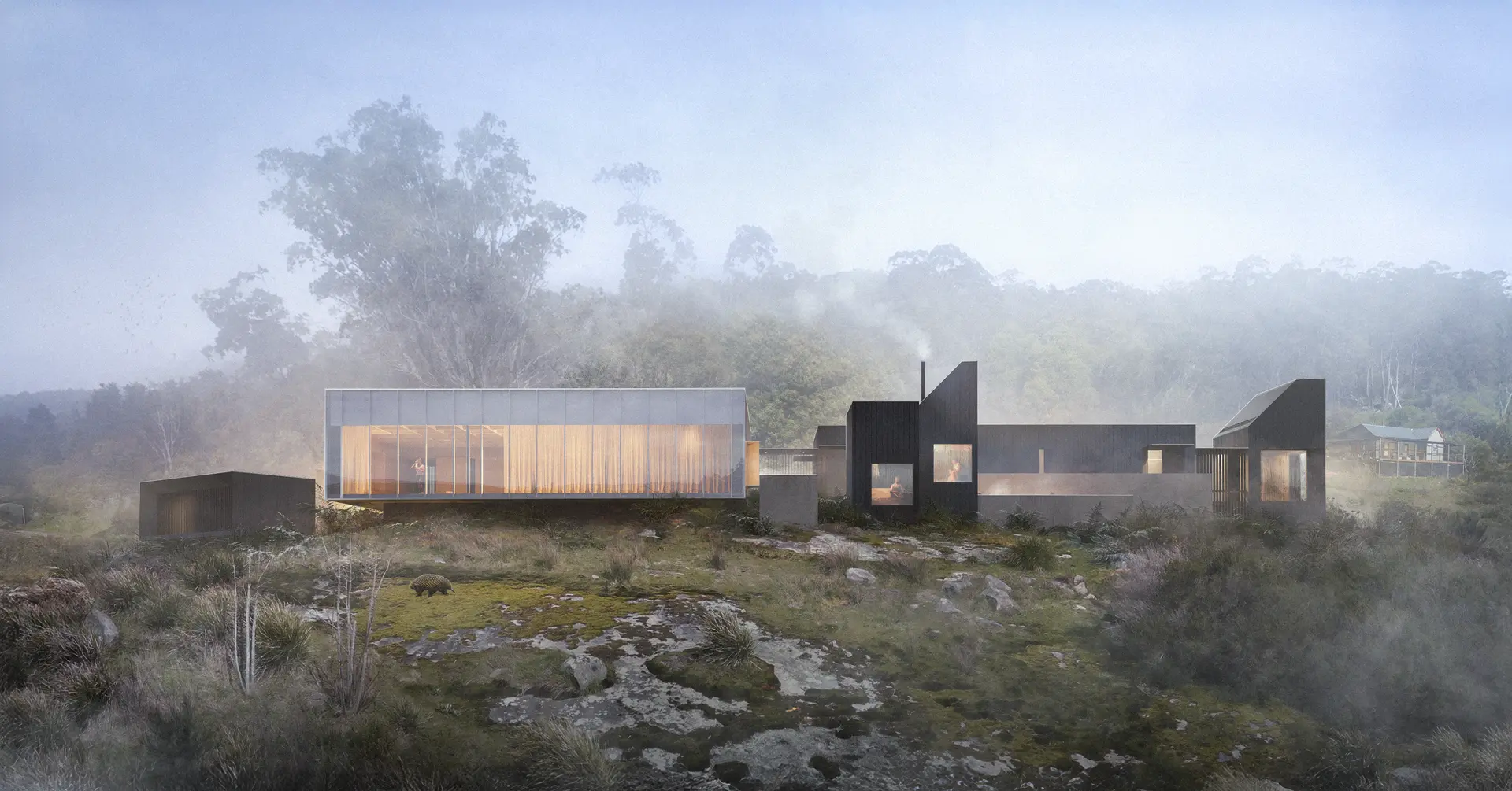 A cluster of low, distinct architectural volumes sits within a misty, sloping landscape, combining a translucent glass and stainless steel yoga pavilion with darker, vertically clad charred timber forms. The composition reads as a series of separate buildings connected by paths, with soft light glowing through the main studio volume, framed by native vegetation and rocky, mossy landscape.