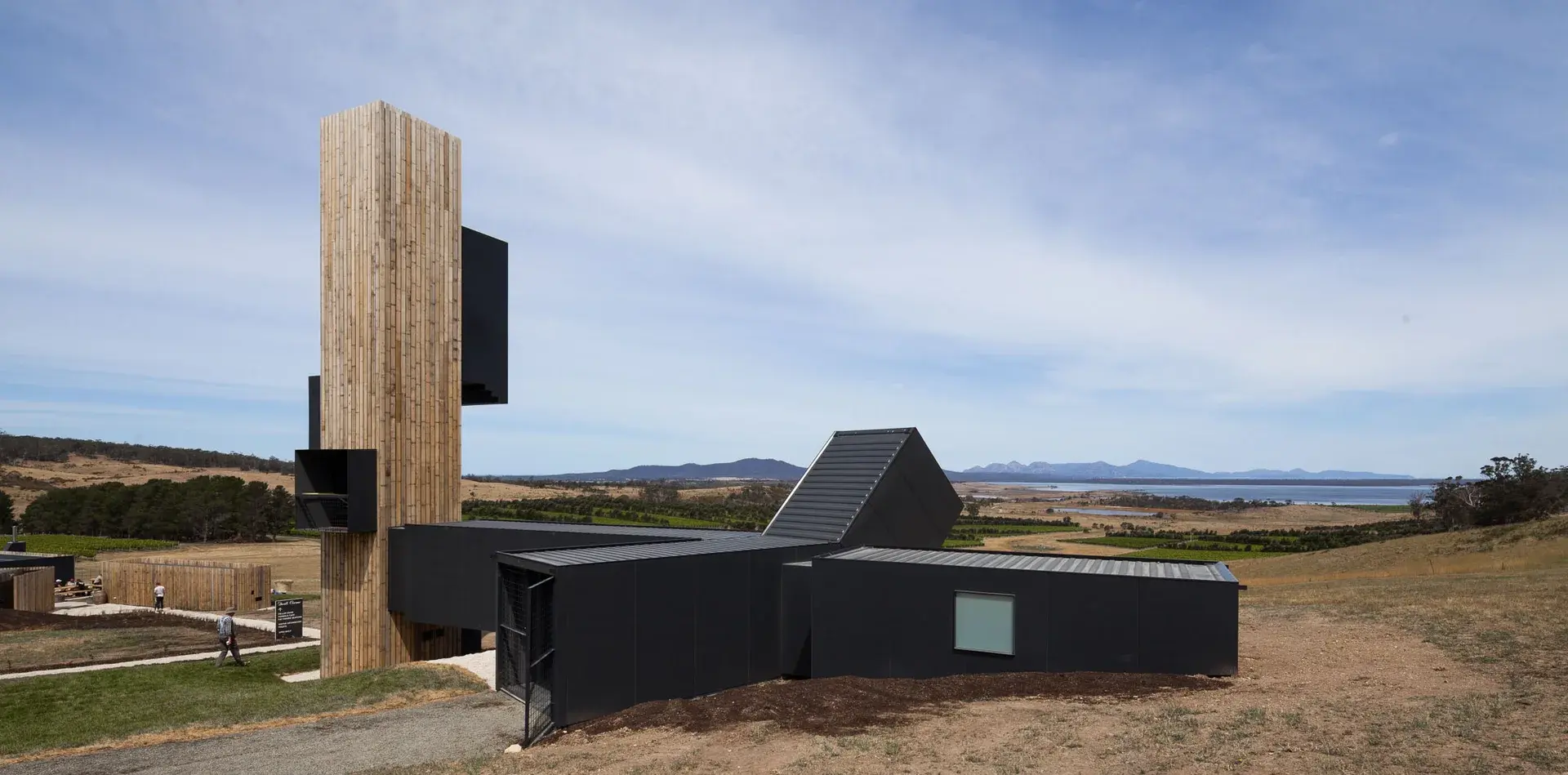 A vertical timber tower and an angular black structure rise from a grassy hillside, overlooking a vast landscape of vineyards and distant mountains.