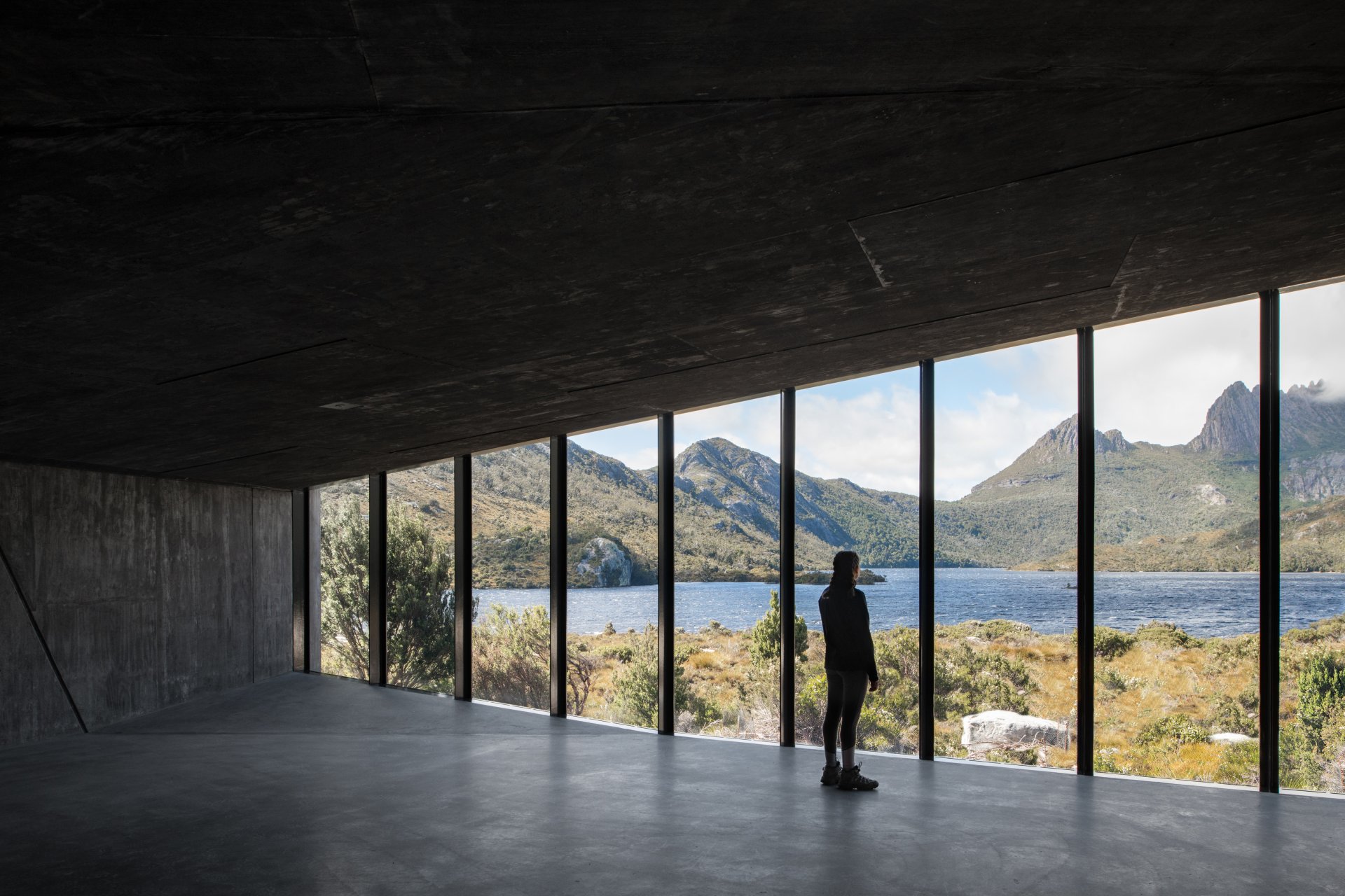 A person stands within a minimalist, dark concrete interior, looking through floor-to-ceiling glass panels at a rugged mountain lake.