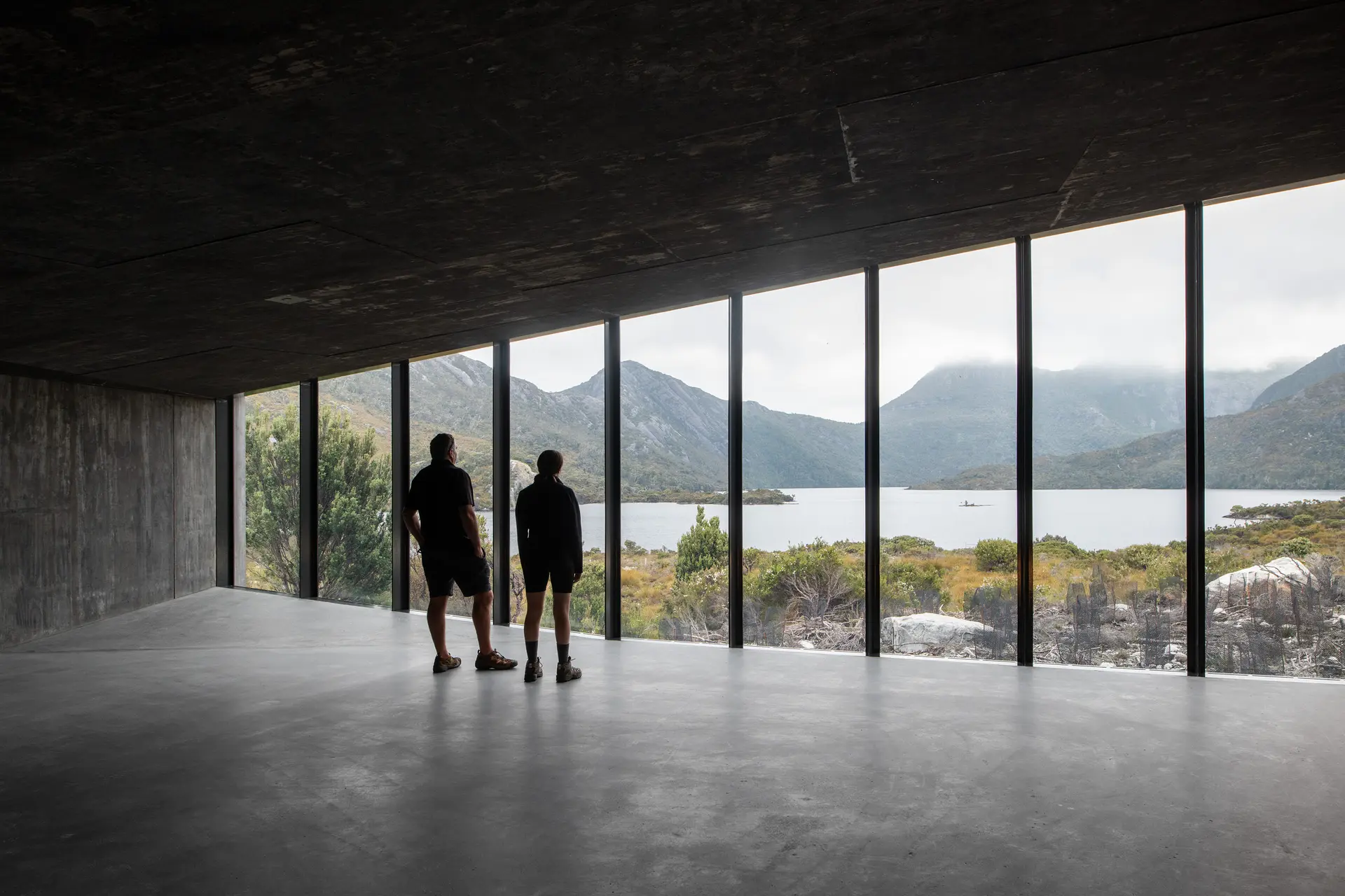 Two people stand in a minimalist, concrete interior, looking out through a wall of floor-to-ceiling windows at a serene lake and rugged, mist-covered mountains.