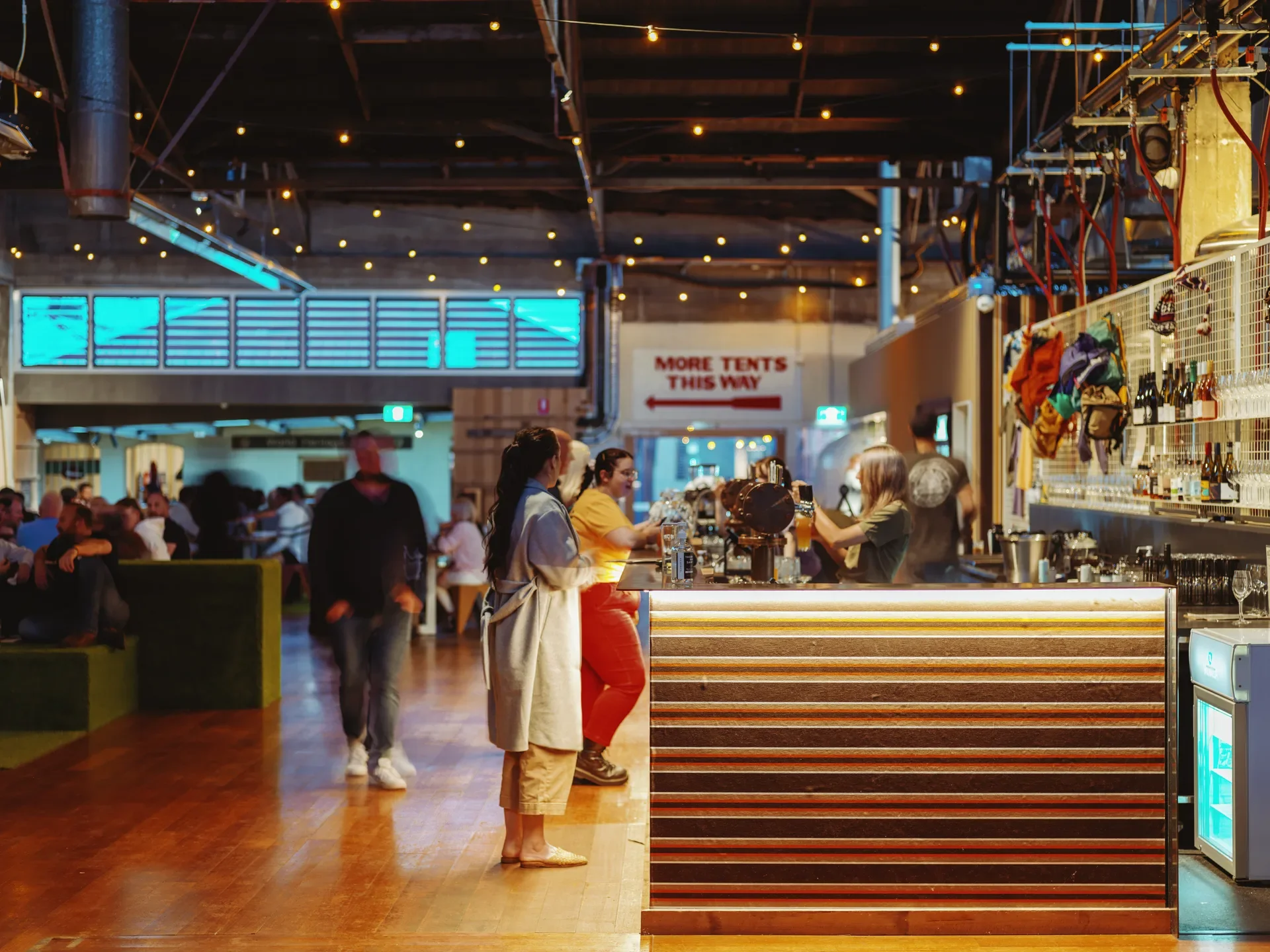 A warm, industrial-style bar features a long, ribbed wooden counter illuminated by a glow from beneath the counter, with people gathered across a polished timber floor. Overhead, the industrial high-ceilinged space is decorated with delicate fairy lights.
