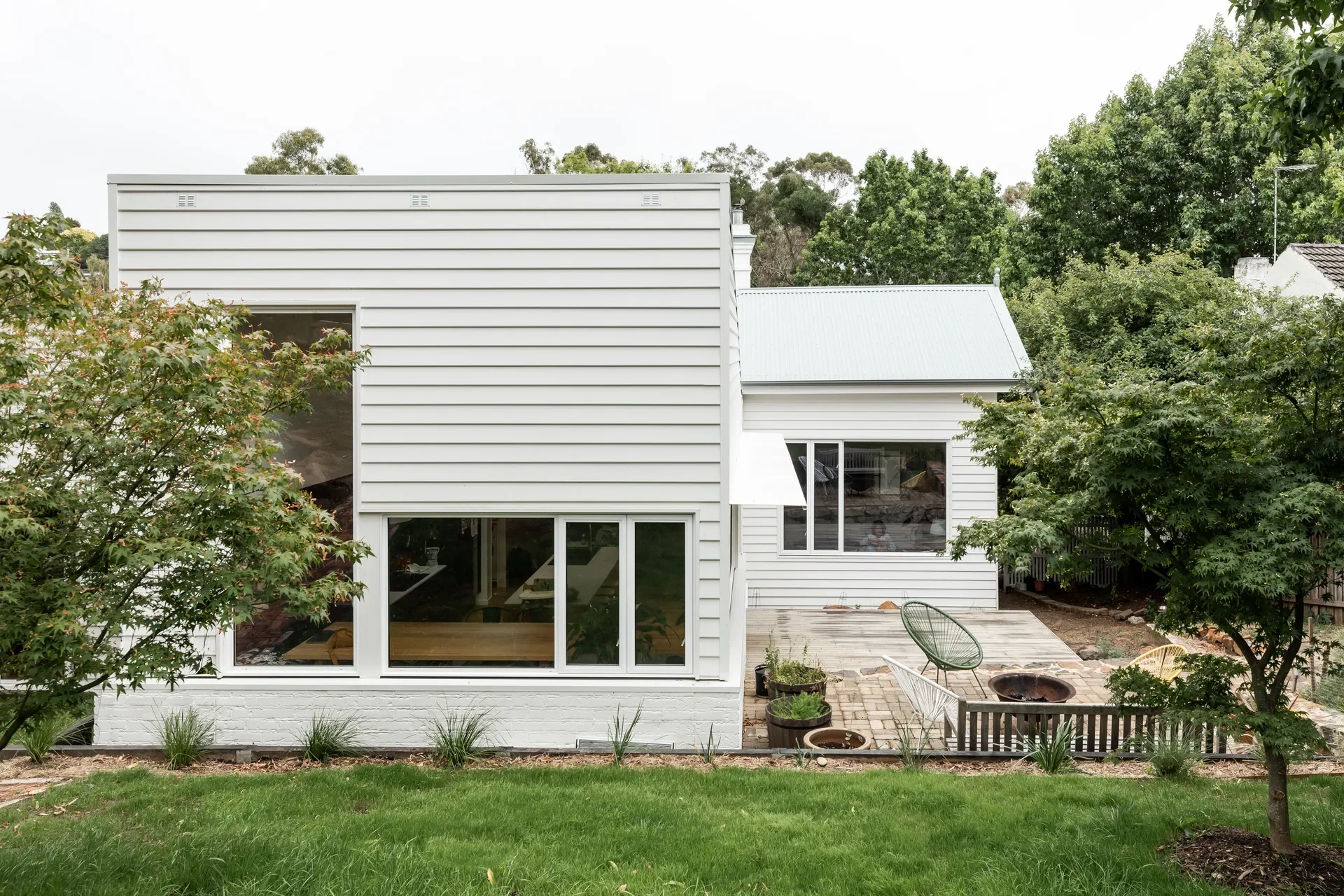 The rear of a white weatherboard house features a modern extension with large glass windows and a lower brick foundation. A paved patio area includes a fire pit, several chairs, and potted plants, all surrounded by lush green trees and a grassy yard.