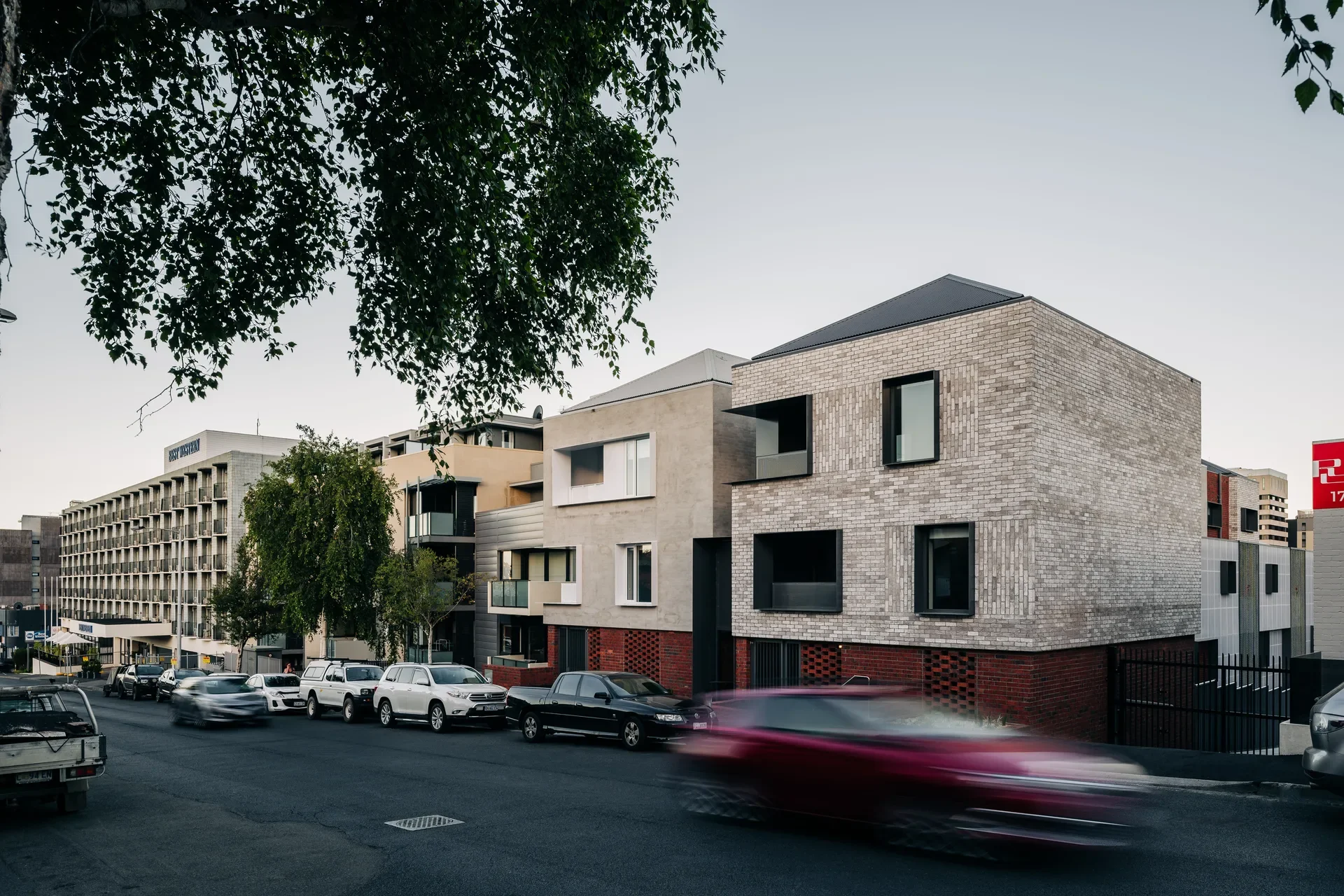 Two modern multi-story buildings both feature a consistent lower strip of red brick, with one finished in a pale brick above and the other a pale render. Both have a pyramid hip roof and deep-set windows standing along a busy street lined with parked cars.