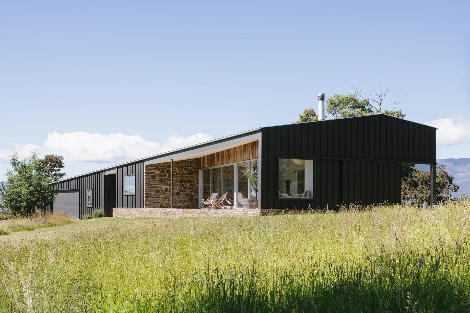 This modern, black vertical-clad home features a long shallow offset open-gable roofline and a stone-walled patio tucked under a timber-lined eave, all set within a field of tall dry grass.