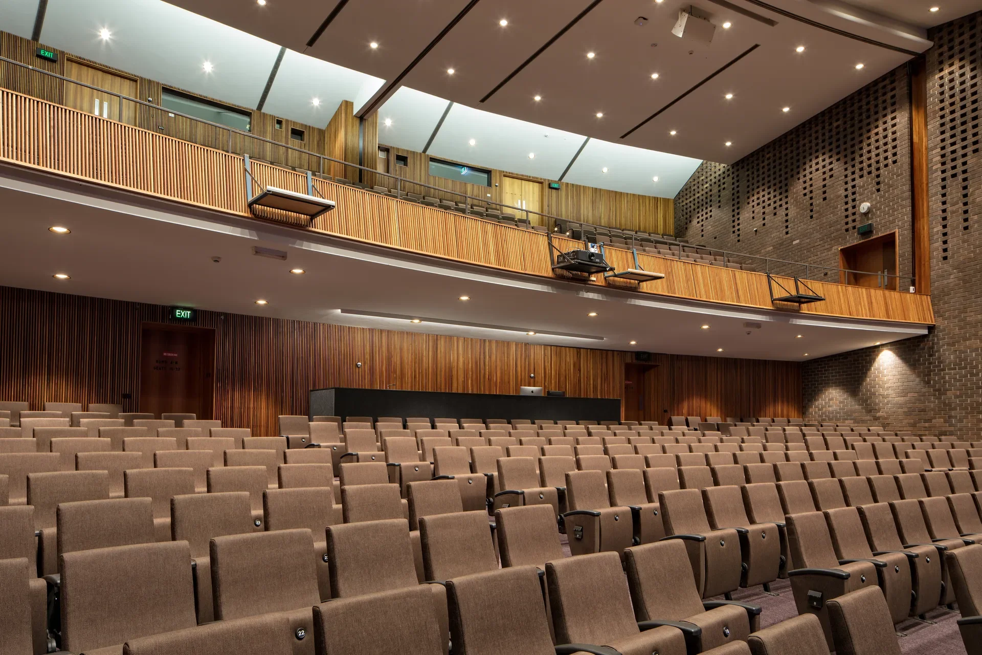 This large, modern lecture theatre features tiered rows of brown upholstered seating. The space is characterised by warm timber-slat wall paneling, a textured brick side wall with decorative patterns, and a prominent wooden balcony level under a ceiling with recessed spotlights.