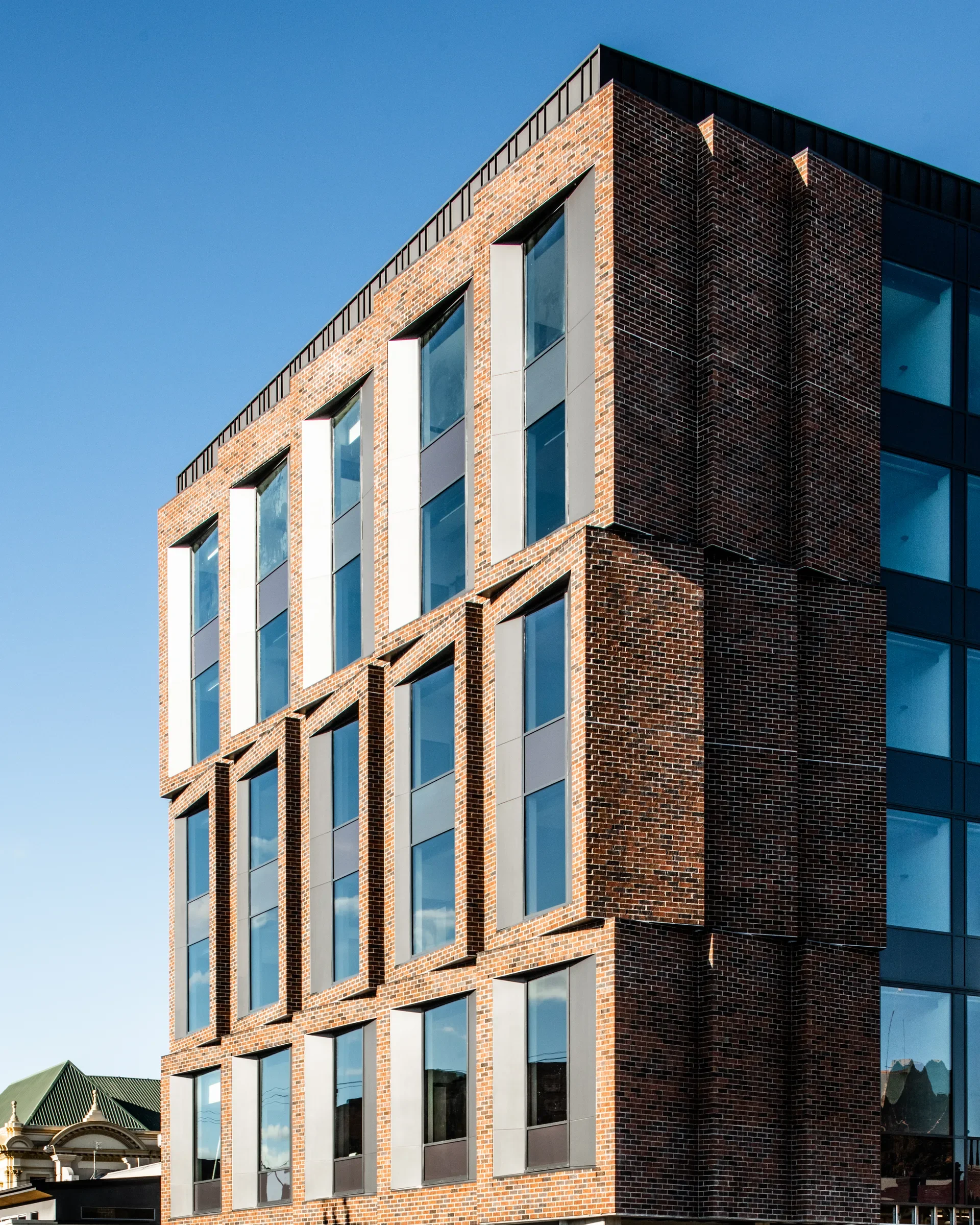 A modern red brick facade features rows of large, deeply recessed windows set at an angle to create a sawtooth-like texture. The windows are framed by grey metal panels and reflect the clear blue sky.