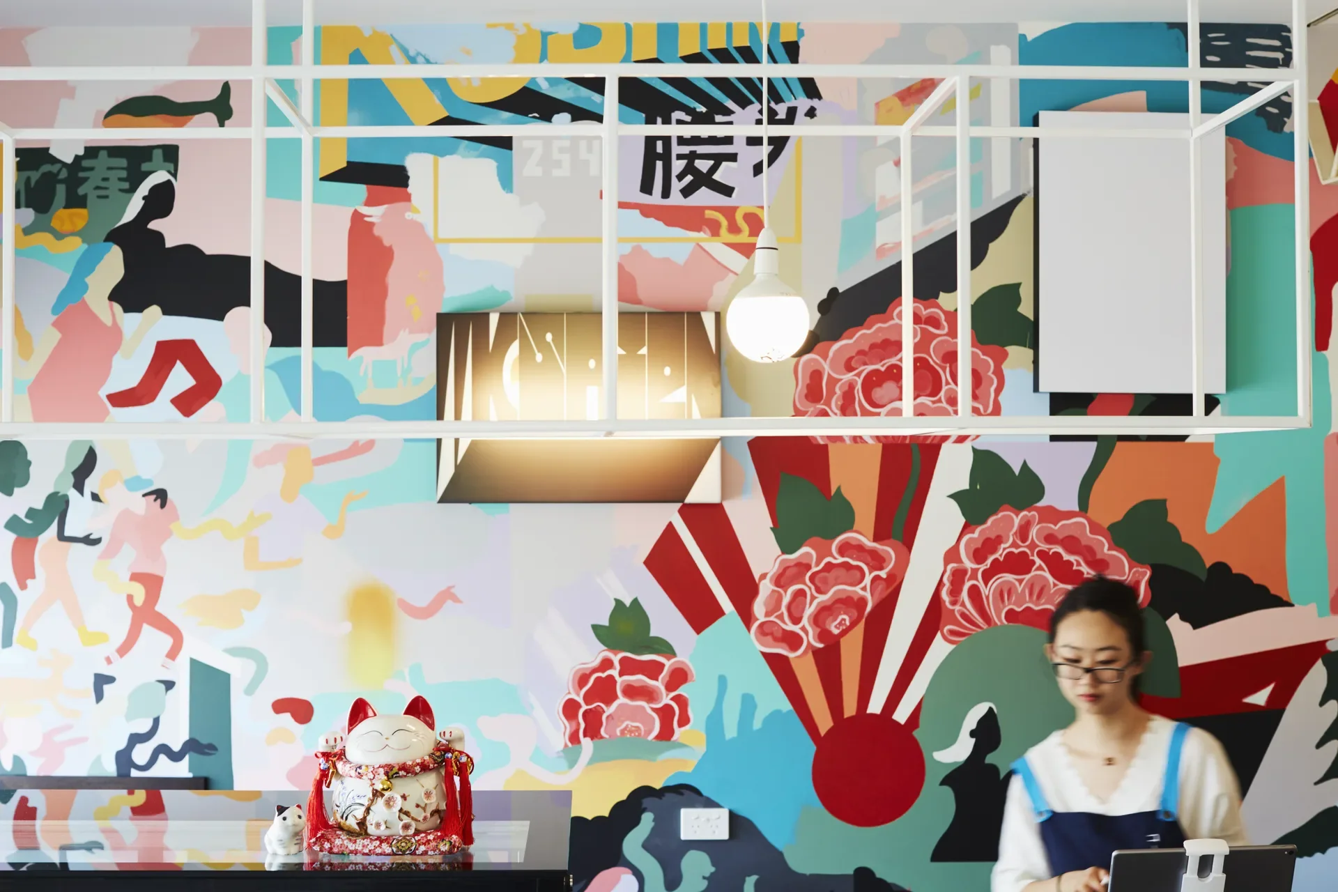 A vibrant, multi-coloured mural featuring floral patterns and Japanese-inspired graphics covers the back wall of this space. A Maneki-neko (lucky cat) figurine sits on a dark counter in the foreground, while a person works with a white geometric ceiling fixture.