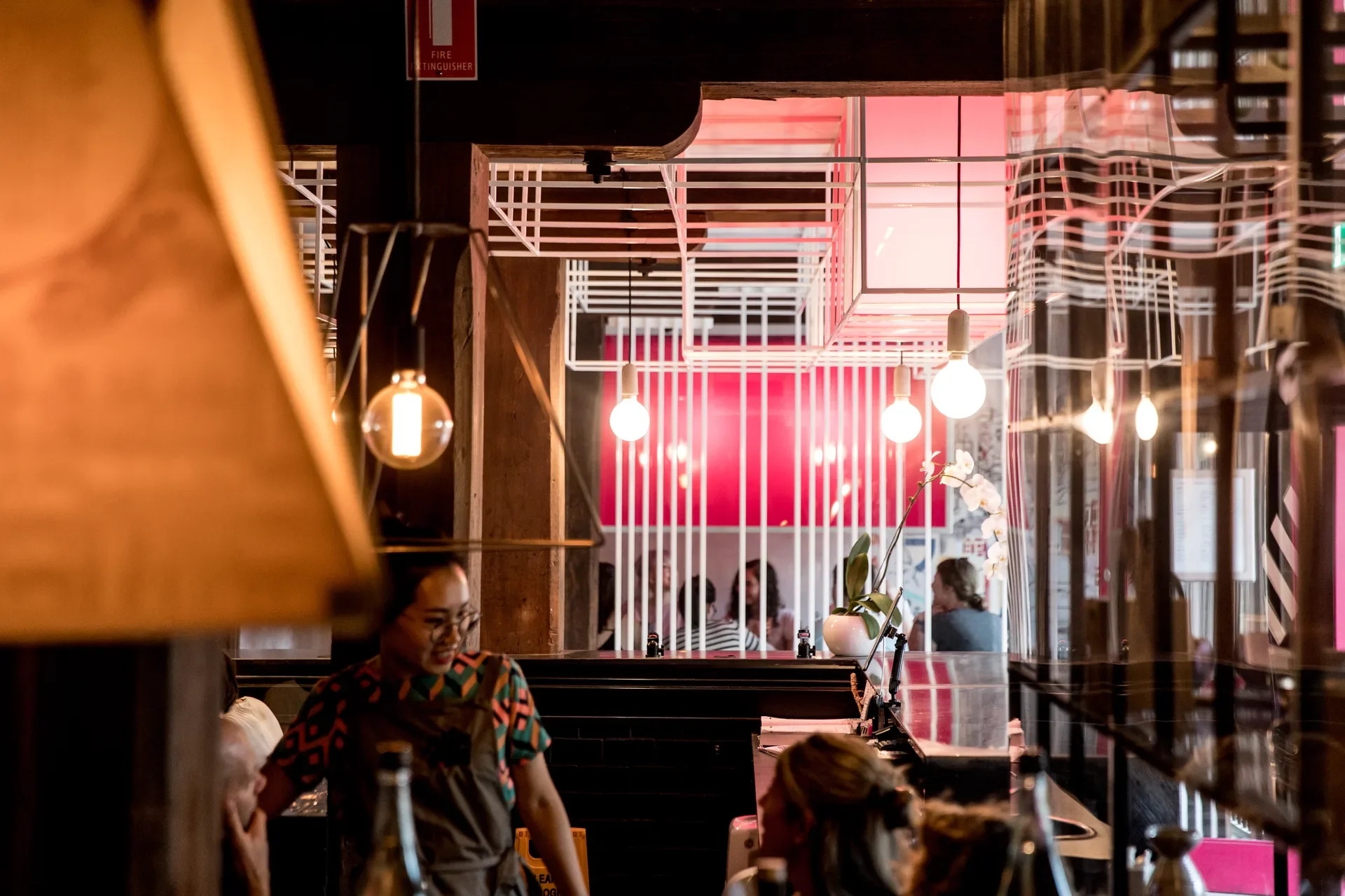 This restaurant interior features pendant light bulbs within a white geometric framework. A vibrant pink wall is visible through white vertical slats behind dining customers.