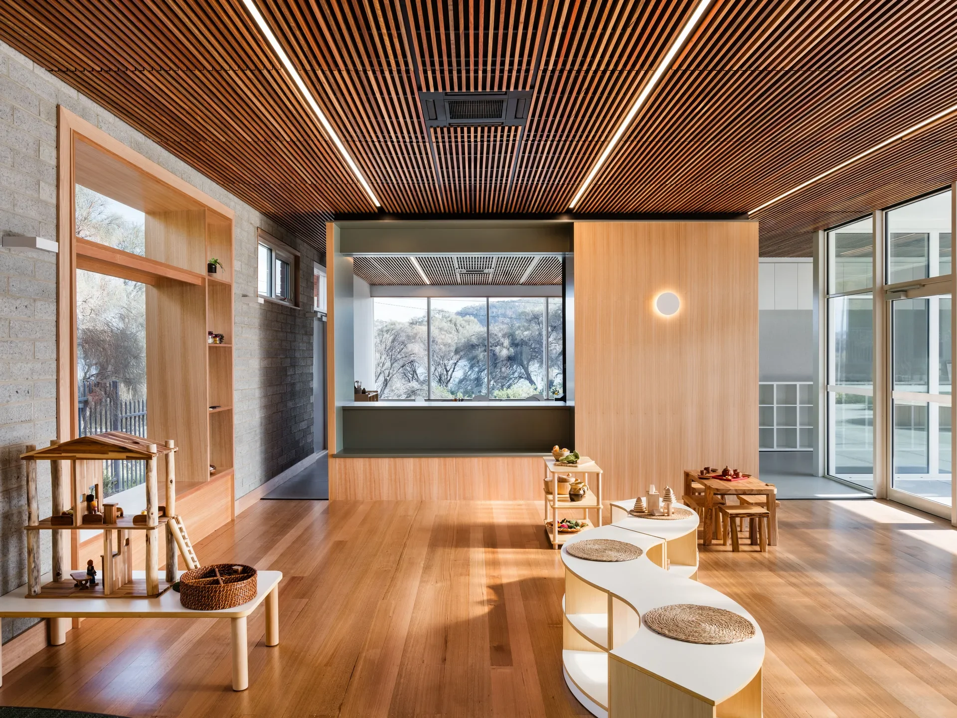 This modern room features slatted wood ceilings, hardwood floors, and light-wood furniture. Large, floor-to-ceiling glass windows and doors provide a clear view of the surrounding trees and landscape.