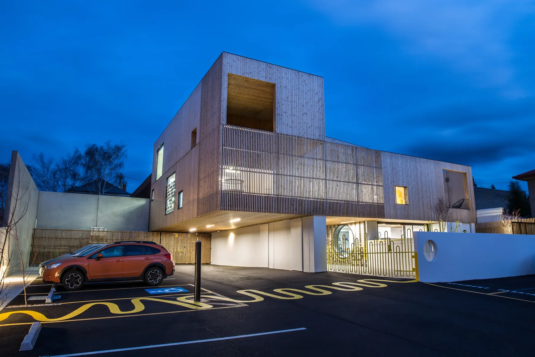 This modern, multi-story building features vertical wood siding and slatted screens. The parking area includes unique yellow wavy line markings that matches the style of fence it leads to, all under an evening sky.