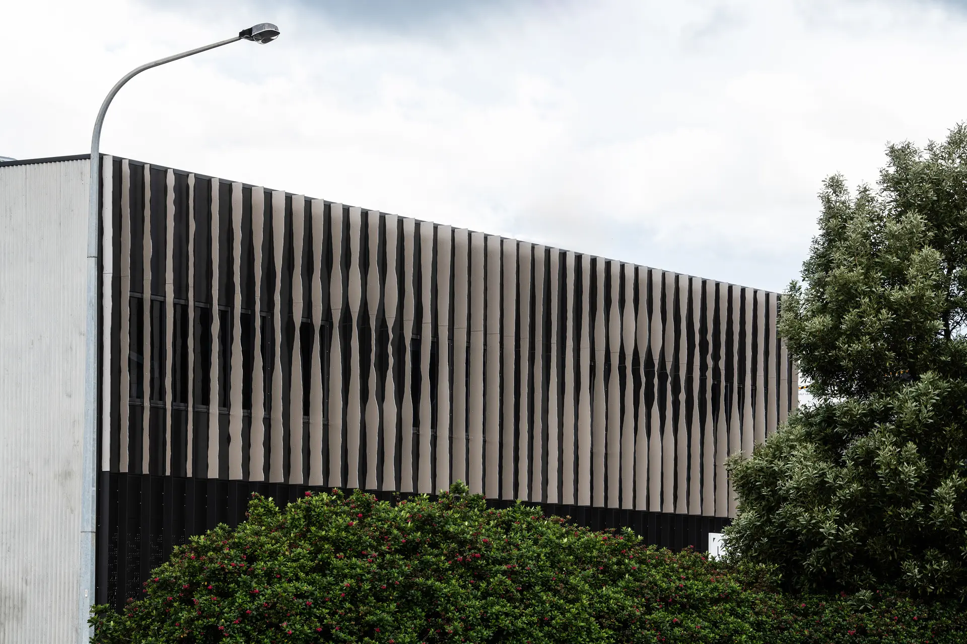 A photograph of the Eye doctors sharp corner condition on an urban street, where a sculpted façade of vertical metal fins wraps the upper level, subtly forming abstracted “eyes” when viewed in motion, while traffic blurs past below and the darker base reveals an undercover carpark recessed into the ground.