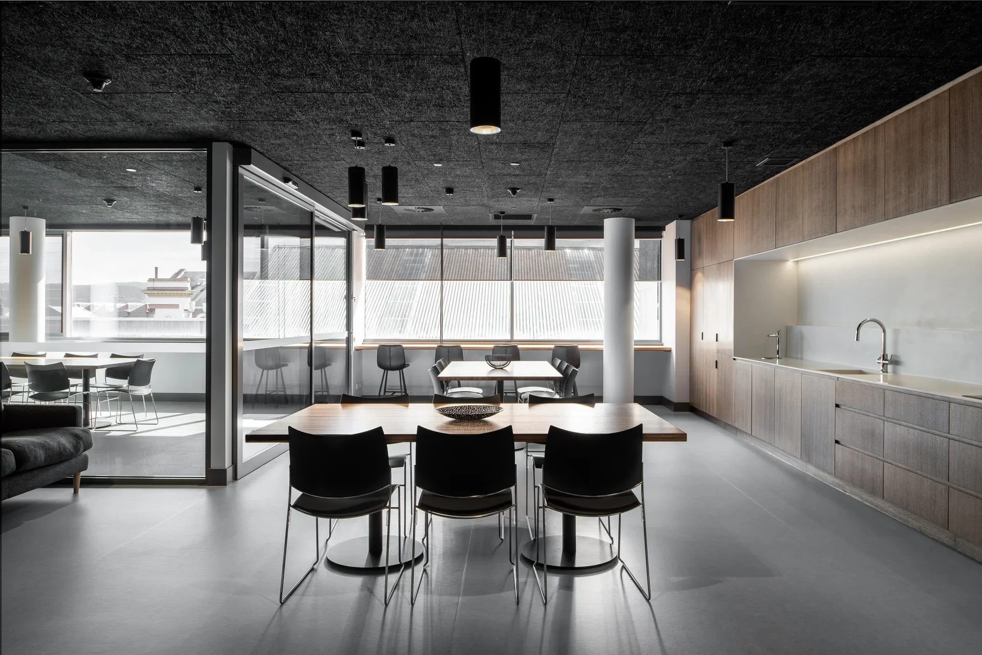 A modern office break room features sleek wooden tables with black chairs under a dark, textured ceiling with cylindrical pendant lights. A long kitchenette with light wood cabinetry and a white backsplash runs along the right wall, while large windows in the background look out onto neighboring rooftops.
