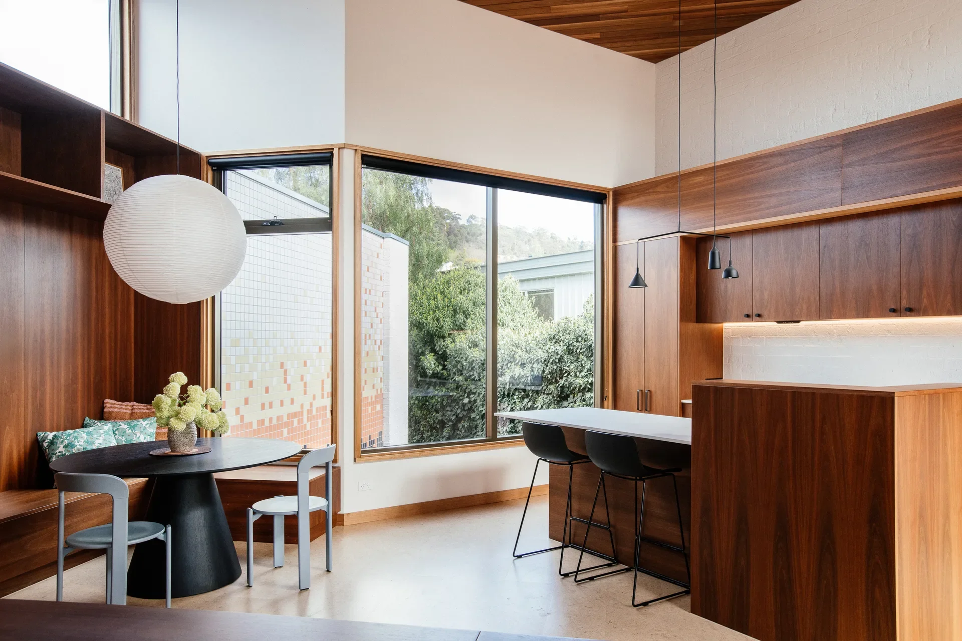 An interior featuring a wood-panelled dining nook and a large paper globe pendant. Large windows in the center overlook lush green trees, filling the kitchen and dining area with bright, natural light.