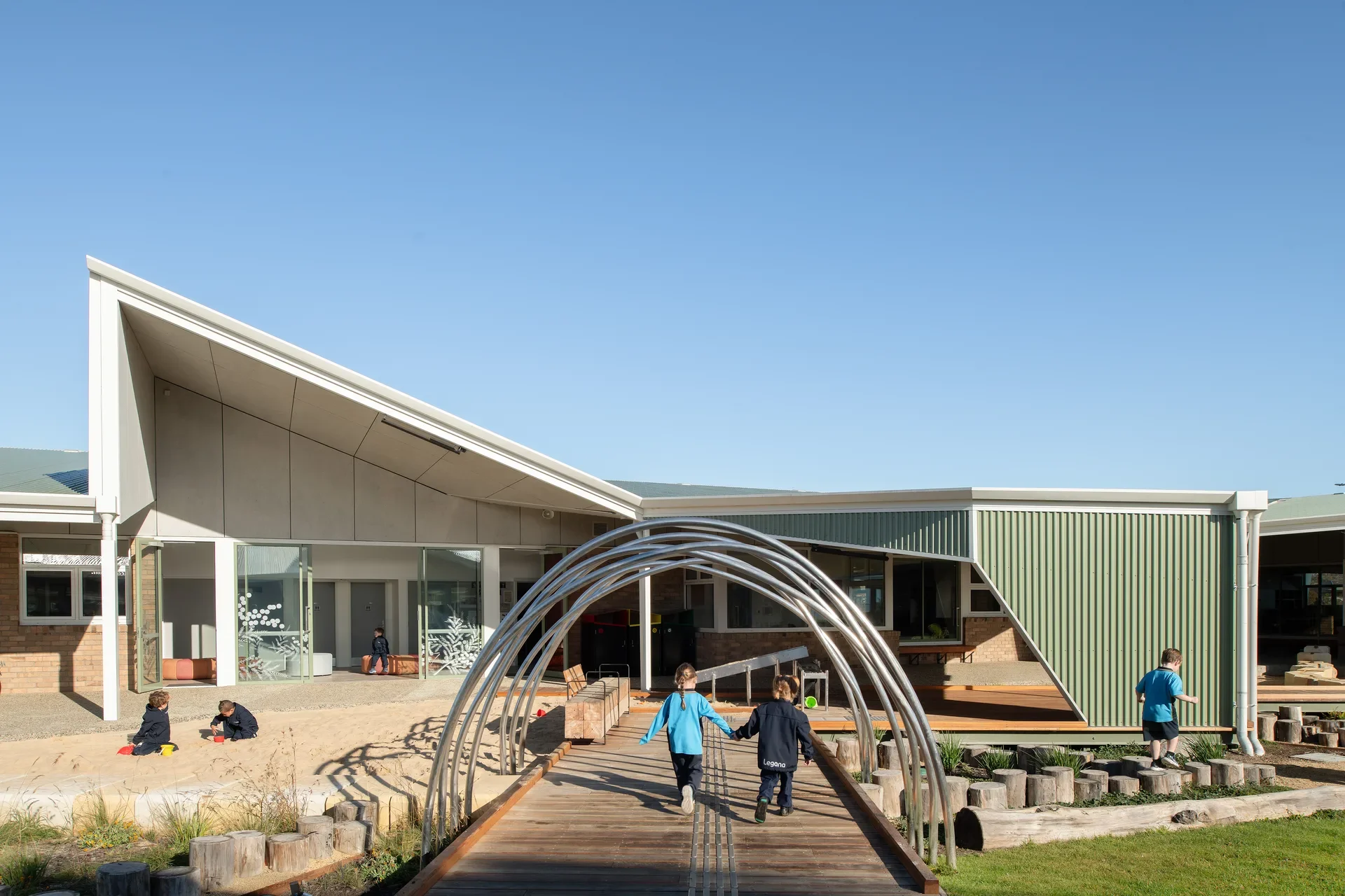 This modern building features a slanted roof and green corrugated metal siding. Two children walk across a wooden bridge with a metallic arched canopy, either side, a large sandpit and a grassy play area.