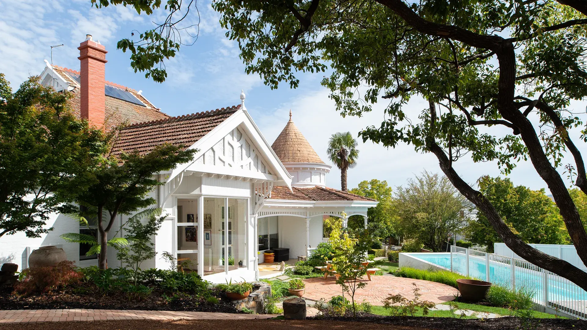 This traditional white house features a gabled roof with decorative timber trim and a unique conical turret in the background. The exterior includes a red brick chimney and large windows that overlook a circular brick patio and a fenced swimming pool surrounded by lush trees.