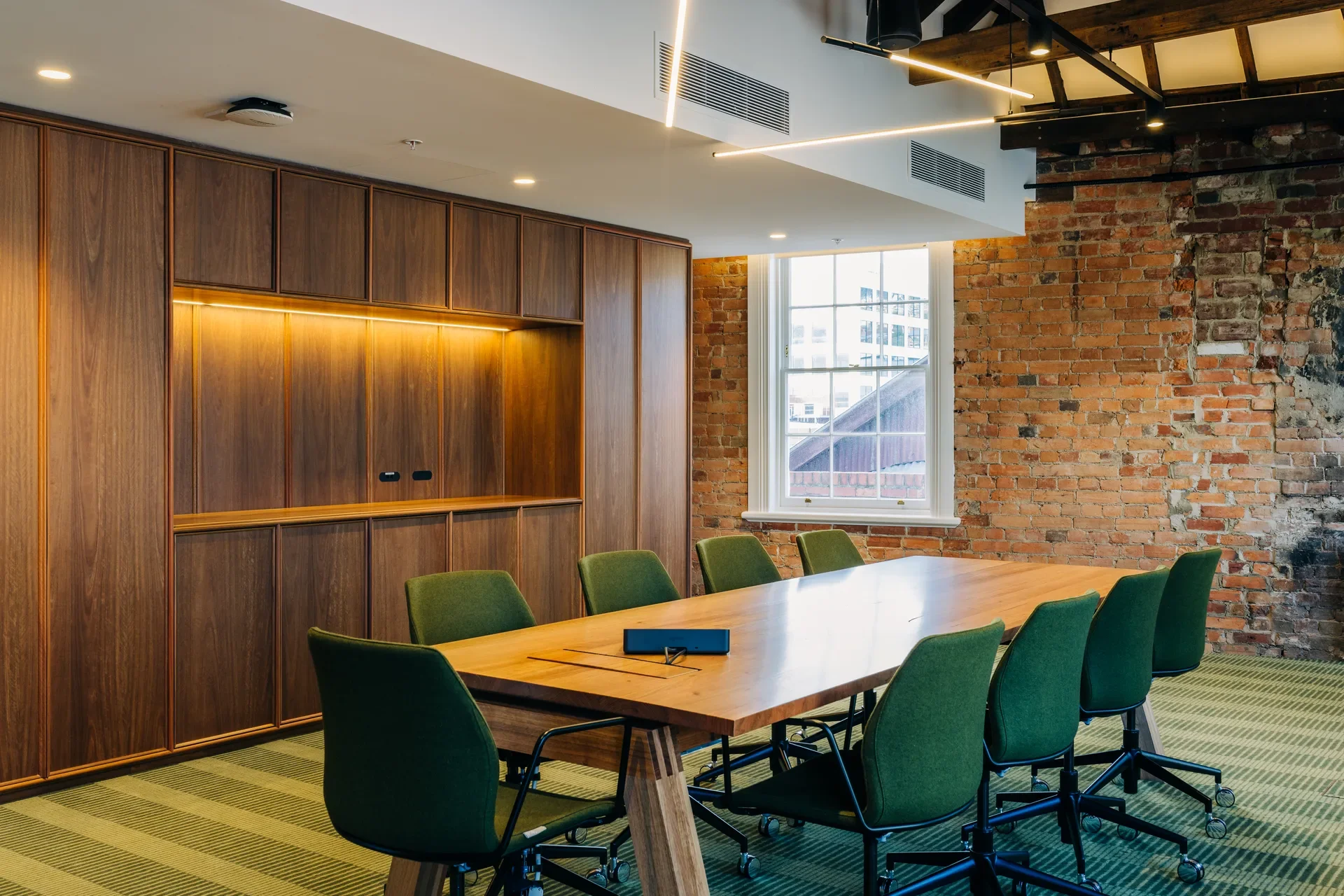 A meeting room featuring a long timber boardroom table and green upholstered chairs. The space blends industrial and modern elements with exposed red brick walls, custom wood cabinetry with integrated lighting, and a linear pendant light overhead.