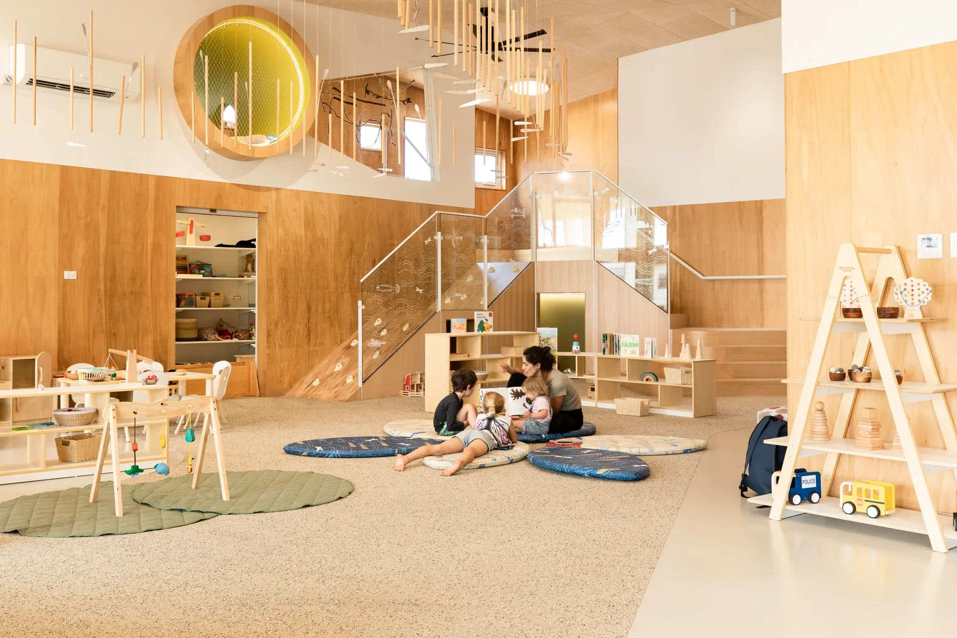 This modern indoor play area features high wood-panelled walls and a large, circular yellow nook recessed high into the wall. An adult sits on flower-shaped floor cushions reading to children near a low wooden bookshelf, while a staircase with etched glass railings leads to an upper level.