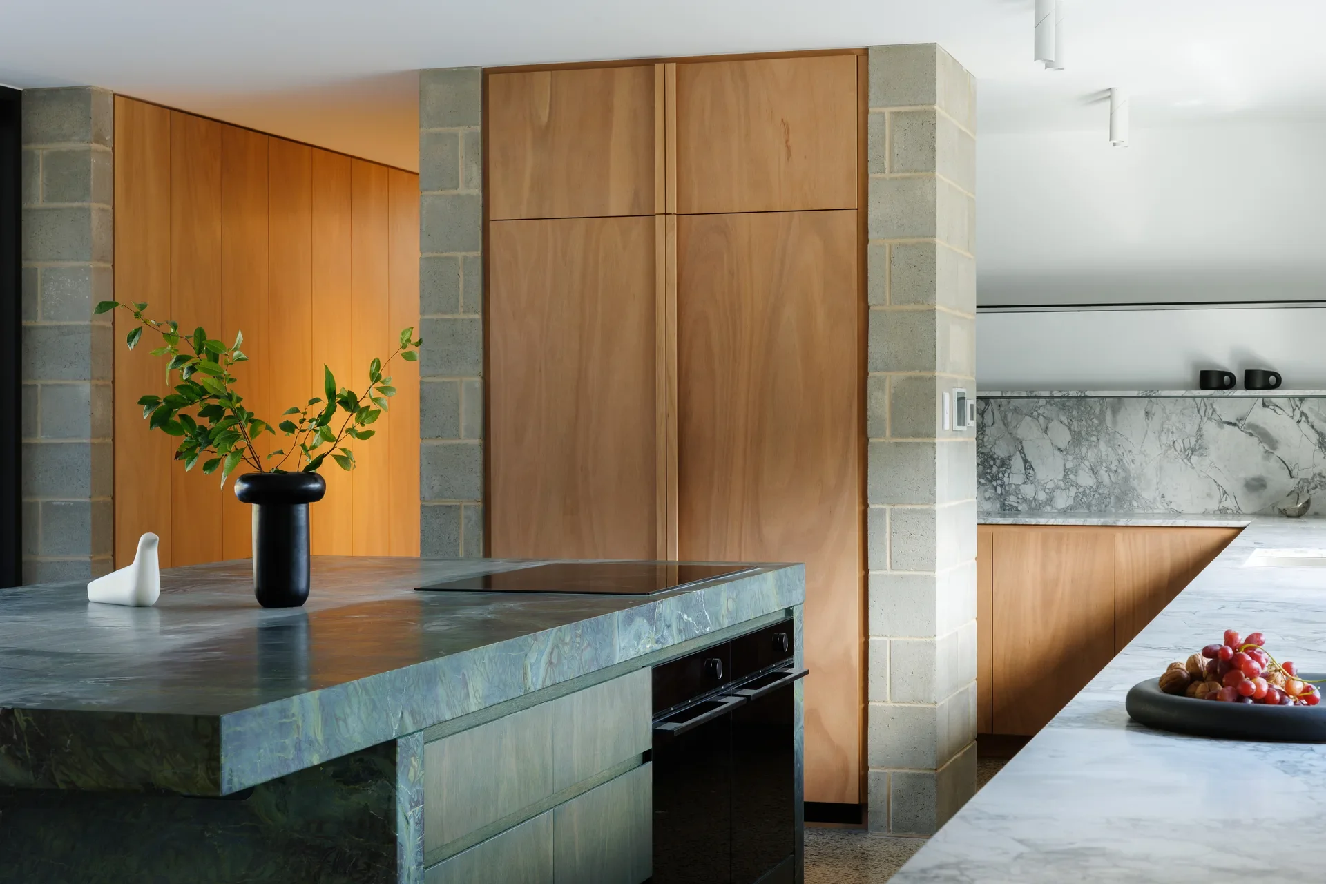 This modern kitchen features a thick green marble island with an integrated black cooktop. The space is framed by a light grey brick pillar and large warm wood cabinetry, with a black vase holding green foliage sitting on the marble surface.