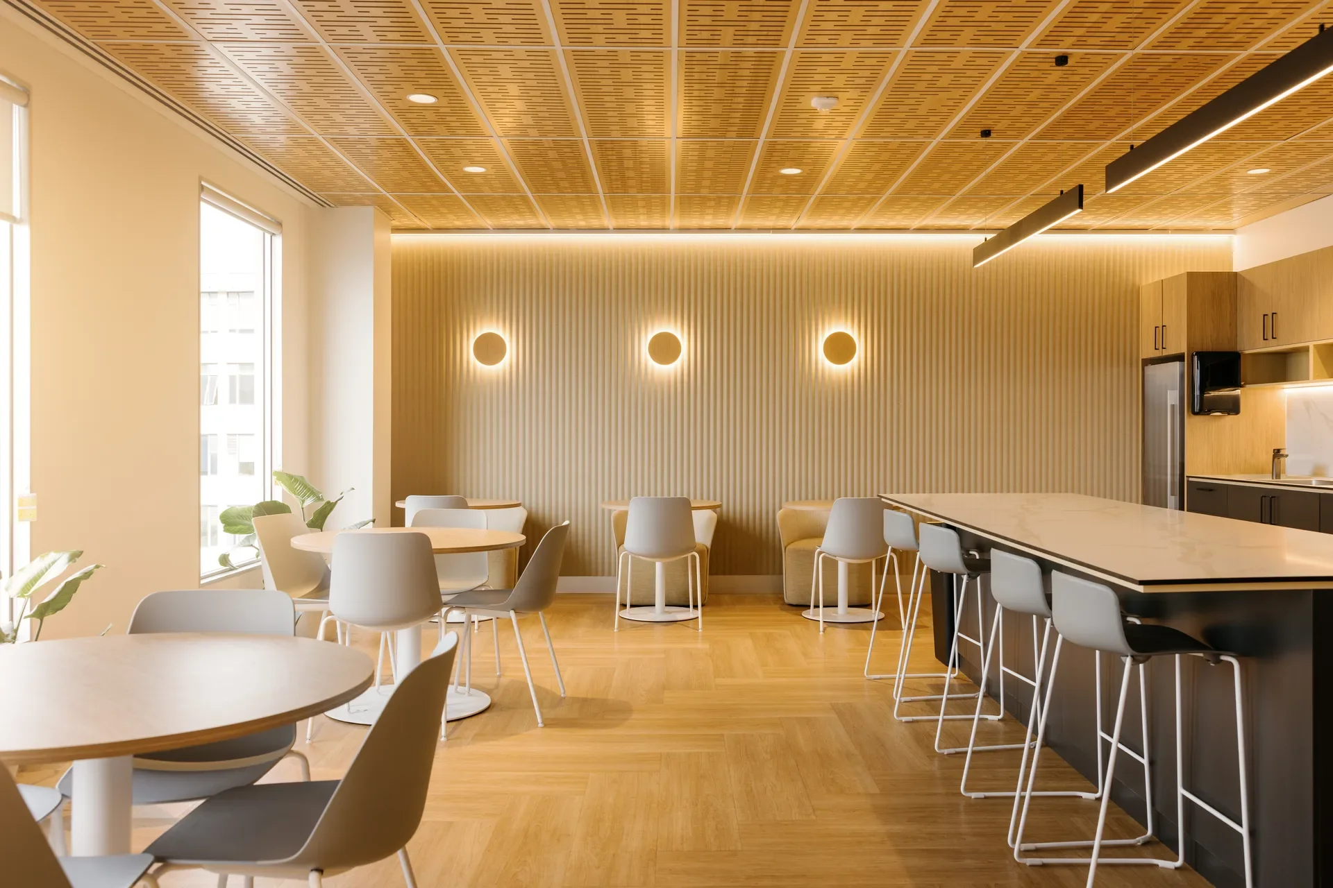 This staff kitchen features vertical-slat wood walls accented by three circular wall sconces and a warm LED light strip. The space is furnished with white and grey chairs at small round tables, beneath a perforated wood-panelled ceiling.