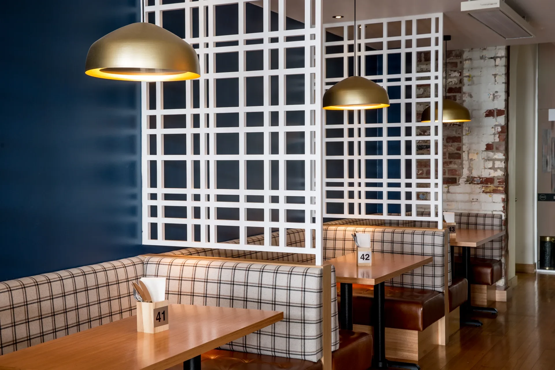 This restaurant interior features plaid-patterned booths with brown leather seats separated by white geometric lattice screens. Brass dome pendant lights hang above wooden tables, while a navy blue wall and exposed brickwork add texture to the space.