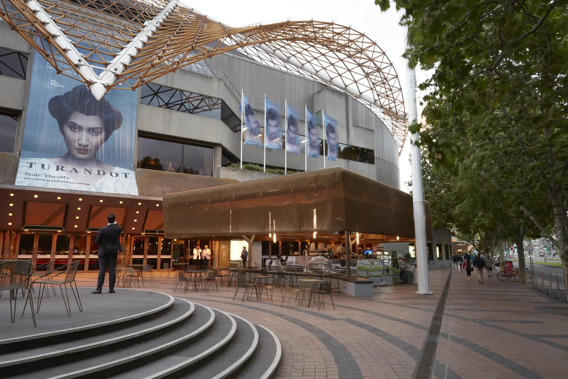 In a paved urban plaza, the Protagonist cafe sits beneath a large, complex geometric metal framework for the "Melbourne Spire". The cafe is partially enclosed by a bronze-mesh screen and features outdoor seating with wire-frame chairs and circular tables.