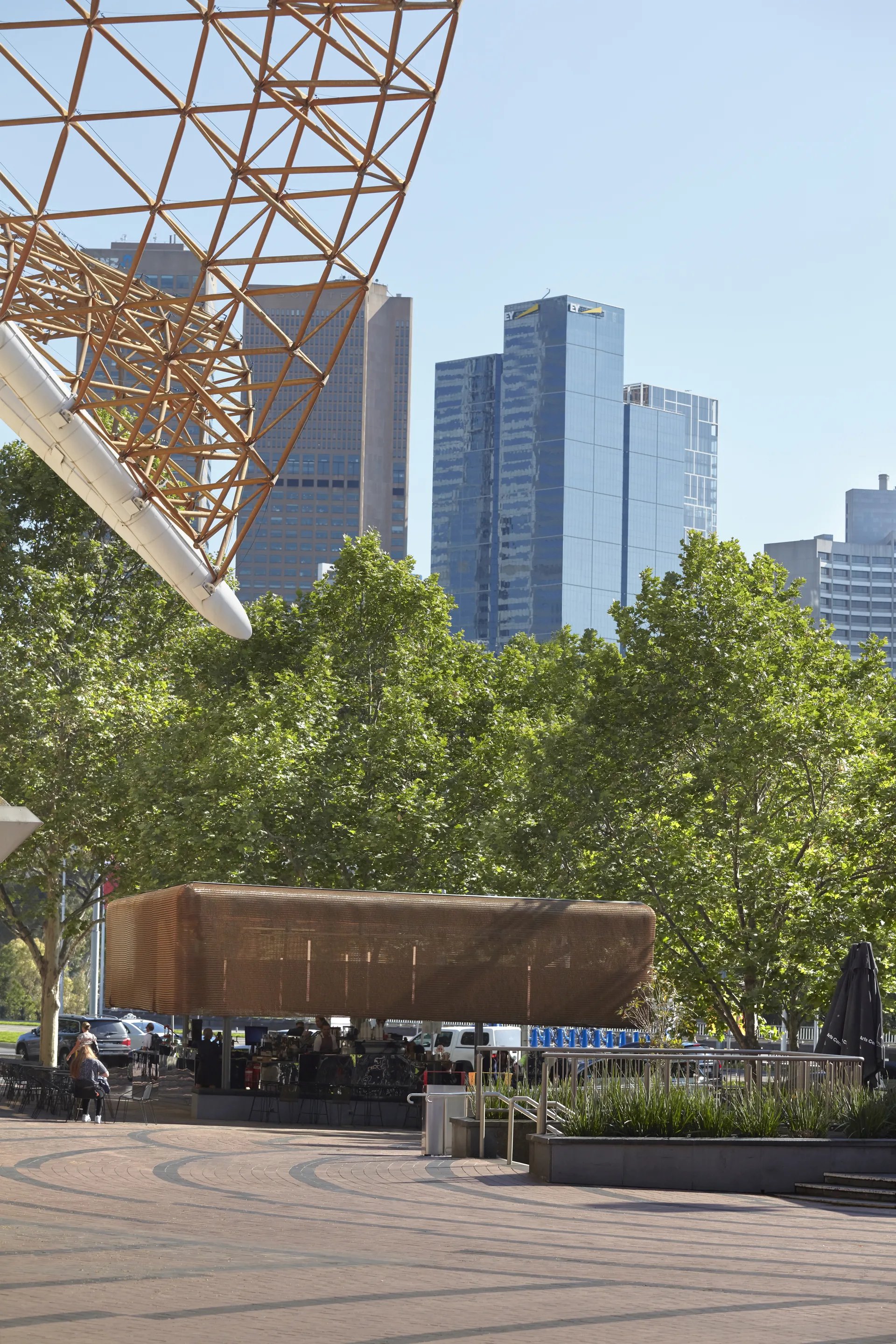 In an urban plaza, a rectangular bronze-mesh pavilion sits nestled among leafy green trees. A complex geometric metal structure frames the upper corner of the view, while modern glass skyscrapers rise in the background under a clear sky.
