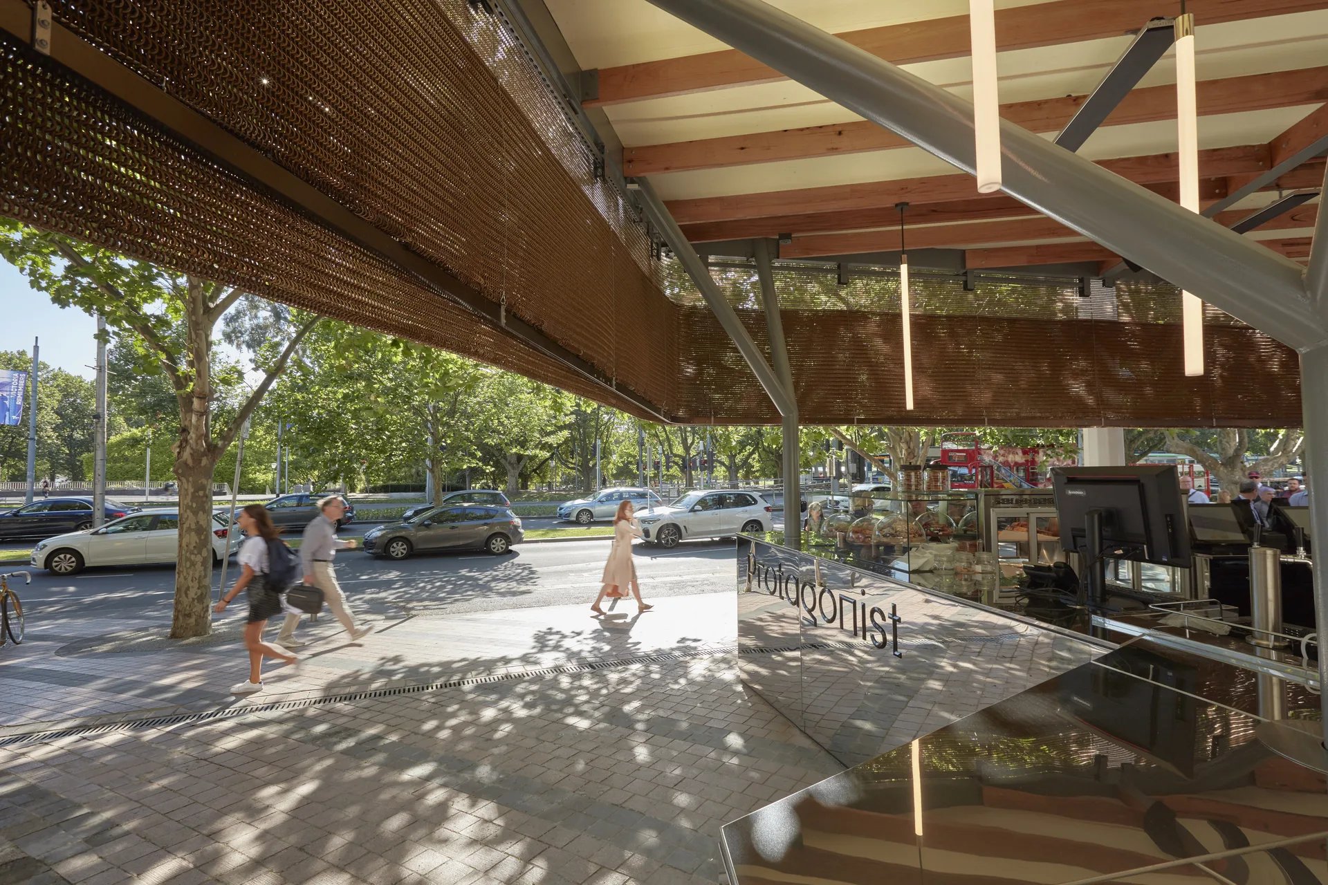 Inside the cafe, a mirrored counter reflects the street while a bronze-mesh screen provides partial shade. The ceiling features exposed timber beams, grey metal supports, and vertical tube lights.