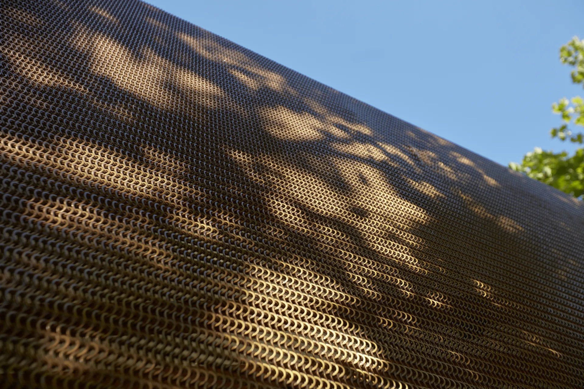 Gemini said A close-up shows the pavilion's bronze mesh, made of small interlocking metal rings. Dappled tree shadows fall across the textured surface against a clear blue sky.