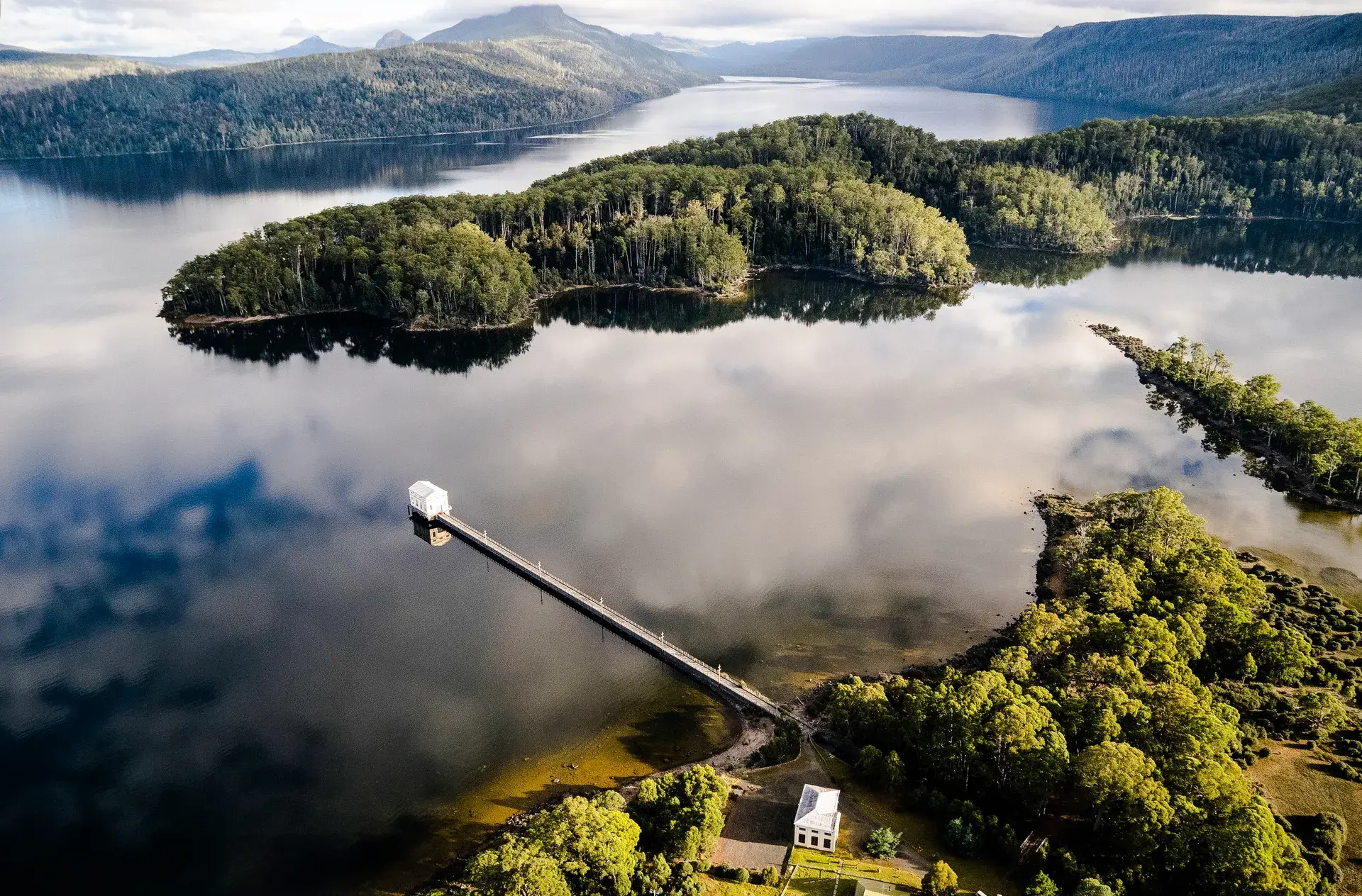 This aerial landscape features a white pump house at the end of a long pier, extending into the still, dark waters of a lake. The water reflects a vast sky of white clouds, while dense green forests and distant mountains surround the shoreline under an overcast sky.