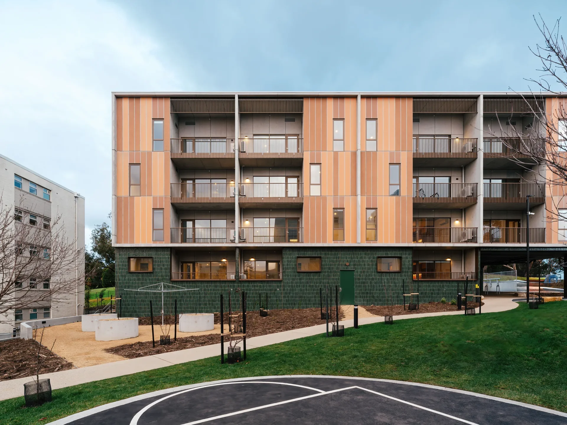 This apartment building features vertical orange and tan panels above a dark green-tiled base. Each unit has a private balcony overlooking a landscaped courtyard with a paved path and a basketball half-court.