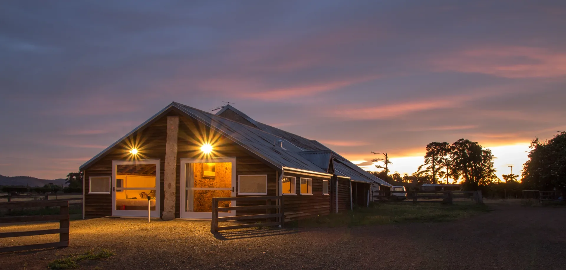 At dusk, a warm light glows from the entrance of a wooden cabin with a slanted metal roof. In the distance, silhouetted trees stand against a vibrant sunset sky filled with pink and orange clouds.