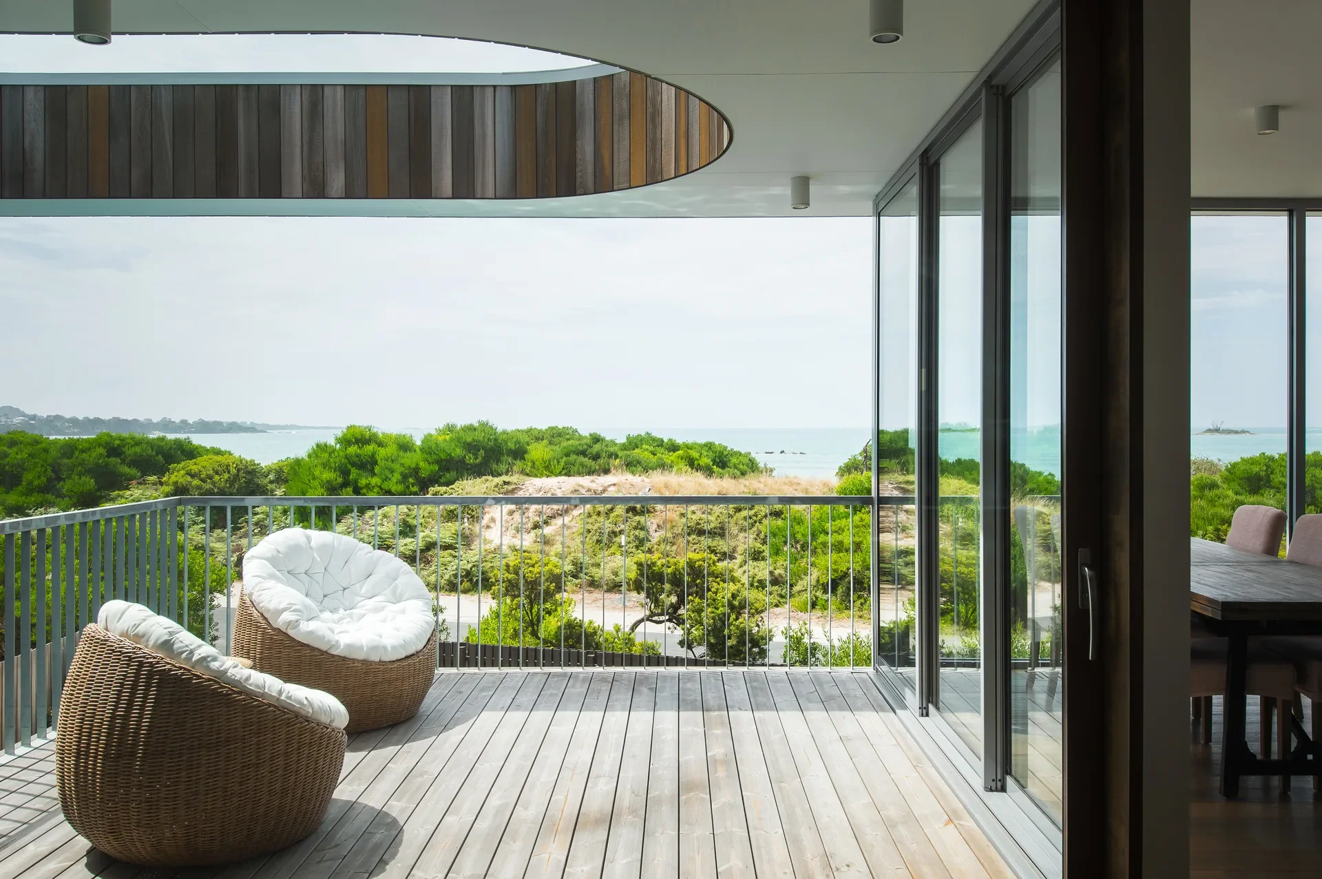 This wooden deck features two round wicker chairs with white cushions, overlooking a lush green landscape and the ocean. Above the deck, a white ceiling includes a unique circular cutout lined with vertical wooden slats.